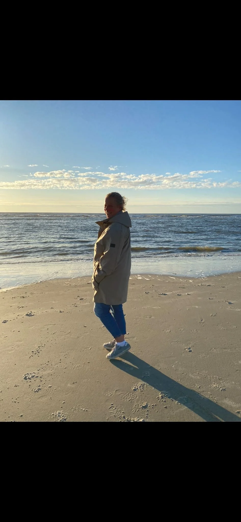 Eine Frau steht am Strand bei Sonnenuntergang, trägt einen hellen Mantel, Jeans und Turnschuhe, während sie in die Kamera lächelt.