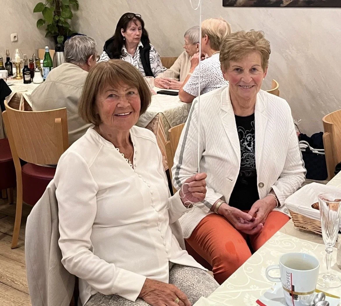 Zwei ältere Frauen sitzen an einem Esstisch in einem Restaurant, eine hält einen kleinen Regenschirm. Im Hintergrund sitzen weitere Personen am Tisch. Es gibt Getränke und Gebäck auf dem Tisch.