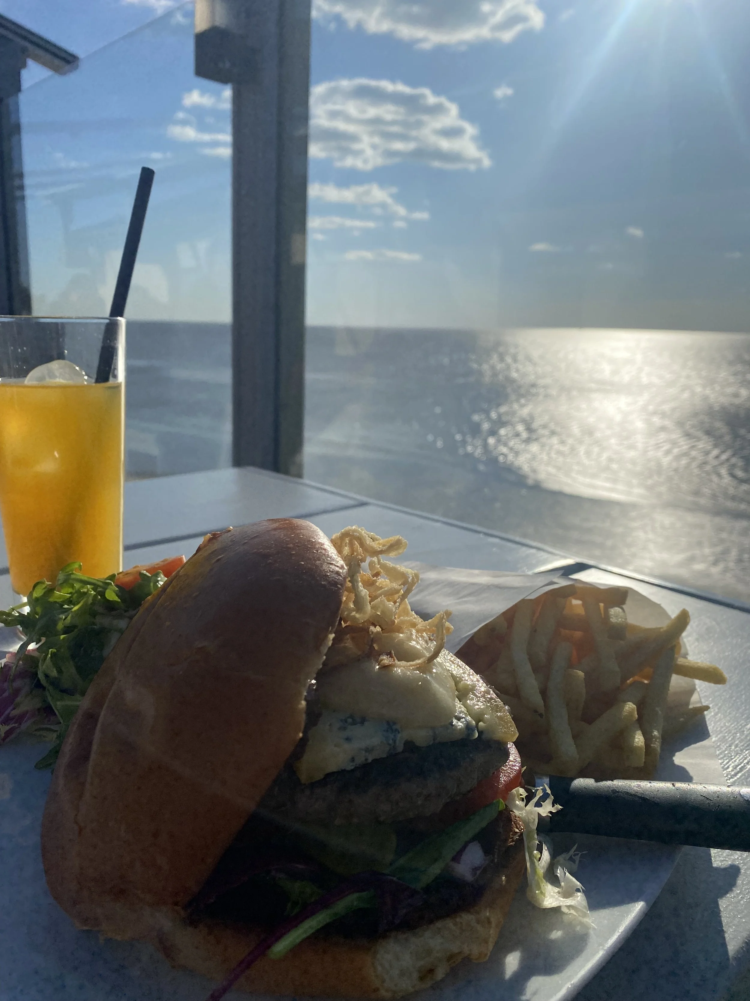 Ein Teller mit einem belegten Brötchen, Kartoffelchips, Salat und einem Glas Orangensaft vor einer Fenster- und Meereskulisse bei sonnigem Himmel.