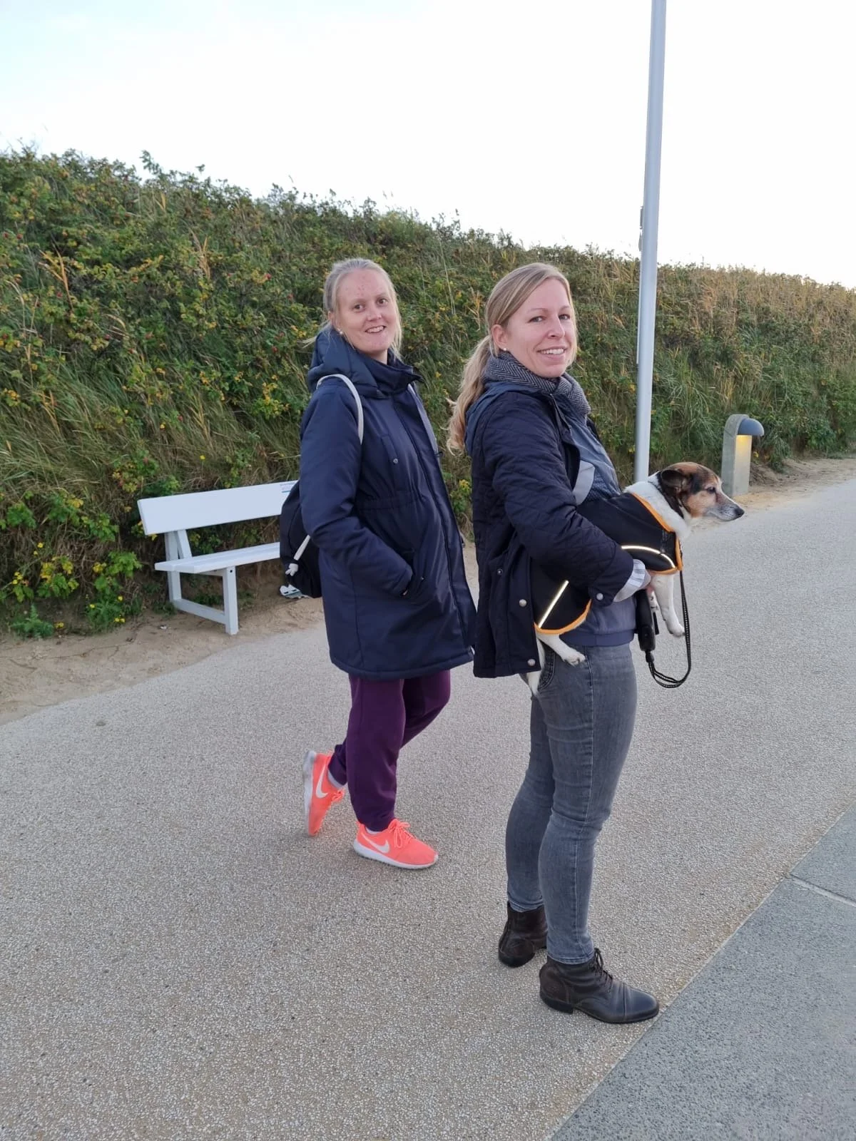 Zwei Frauen stehen an einem Weg, eine mit einem Blindenhund in einer Tasche. Sie tragen dunkle Jacken, die andere trägt lila Hosen und helle Schuhe. Im Hintergrund sind Büsche und eine Bank.