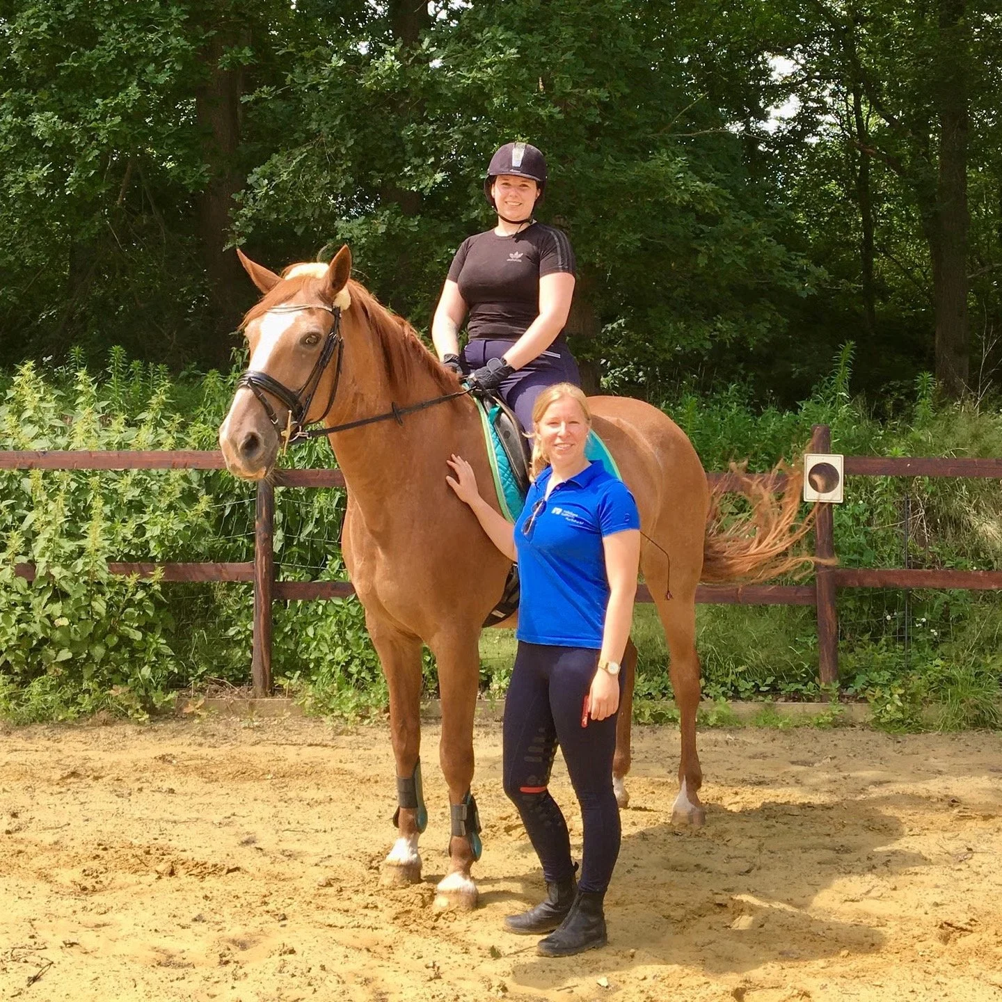 Zwei Frauen und ein brauner Pferd auf einem Reitplatz, eine Frau sitzt auf dem Pferd und trägt einen Reithelm, die andere steht neben dem Pferd in einer blauen Sportjacke.