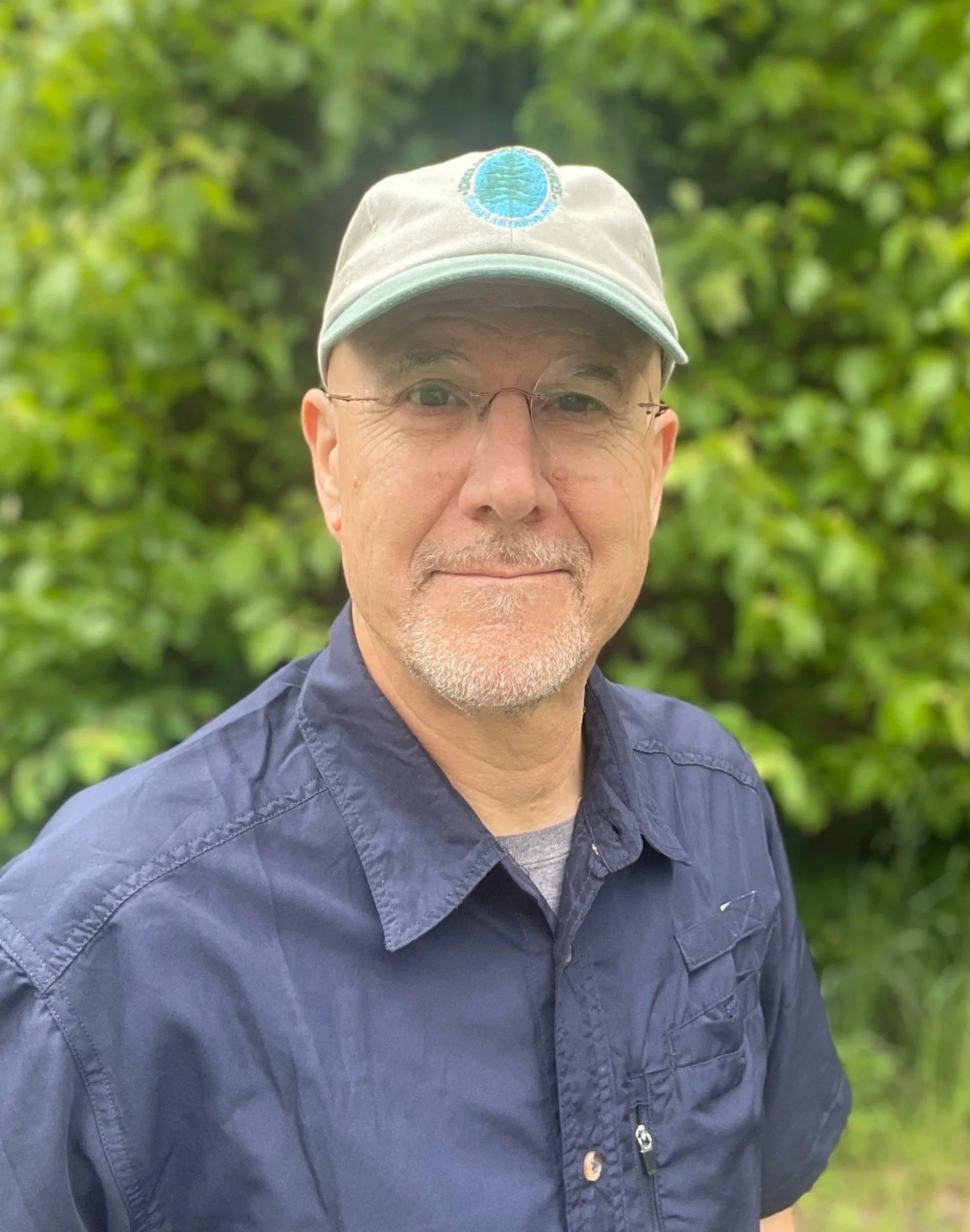 Close-up of a smiling middle-aged man with glasses, wearing a beige cap with a blue logo, and a navy blue shirt, outdoors with lush green foliage in the background.