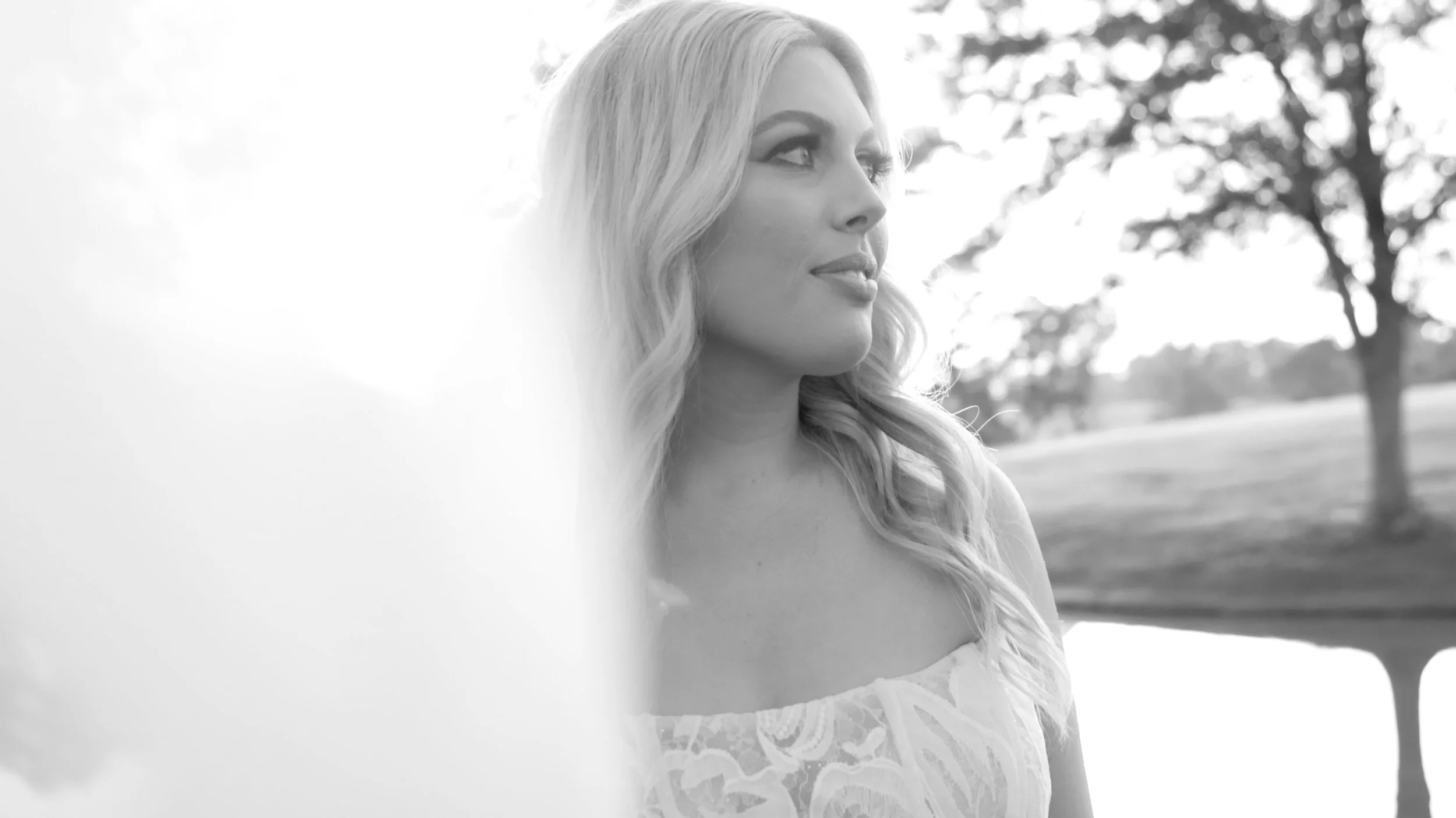 A woman with long wavy blonde hair looking to her left outdoors in a park or garden setting with trees in the background, in black and white