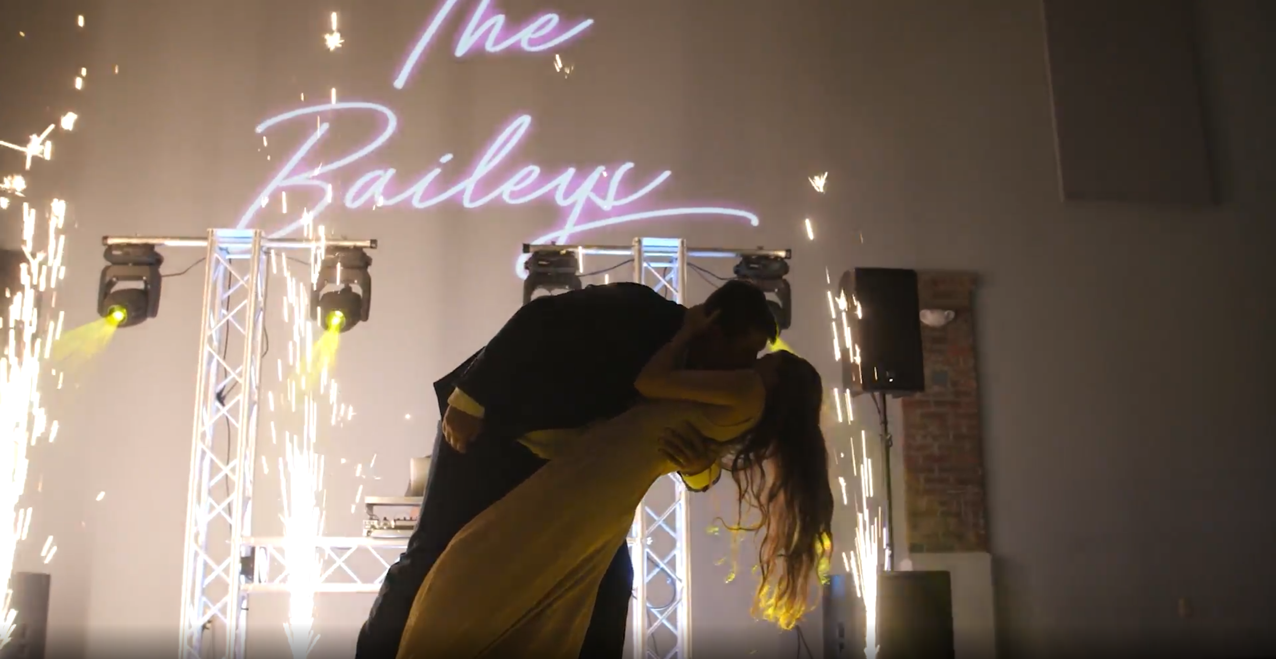 A couple kissing during their wedding reception with sparklers and a neon sign that reads 'The Bailes' in the background.