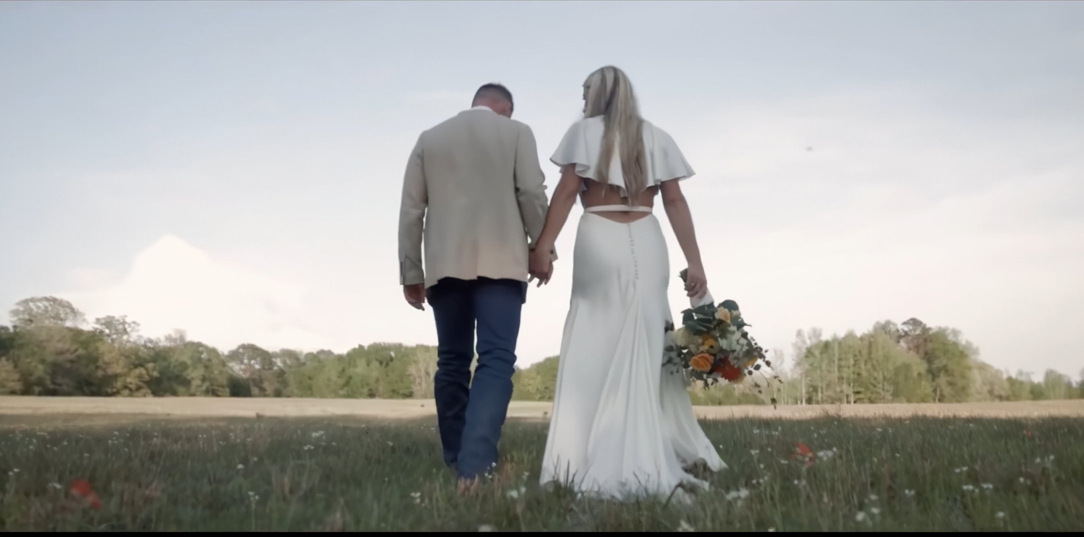 A couple walking hand in hand outdoors during their wedding, with the bride holding a bouquet of flowers, in a grassy field with trees in the background under a cloudy sky.
