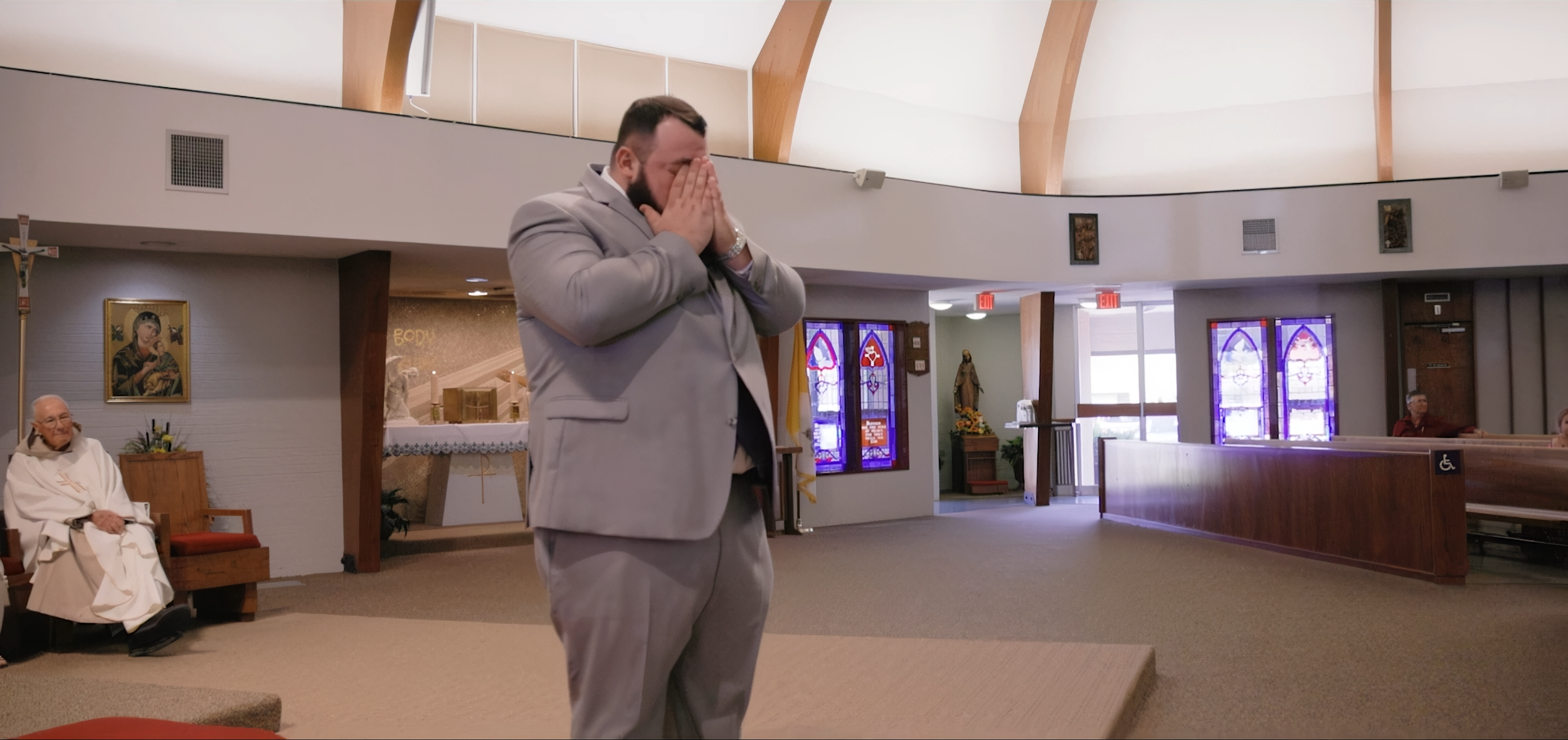 A man in a gray suit with a beard praying with his hands covering his face inside a church near an elderly priest sitting in a chair. The church has stained glass windows and religious artwork.