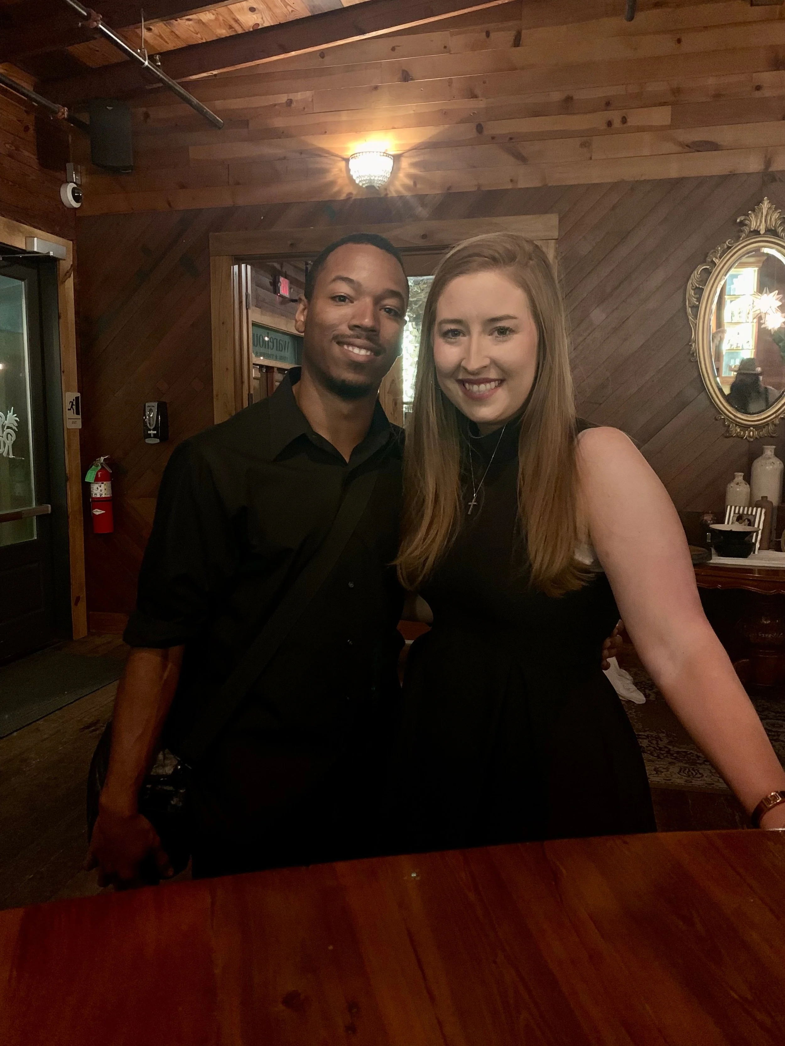 A man and woman pose together indoors, with wooden walls, a mirror, and decorative items in the background.