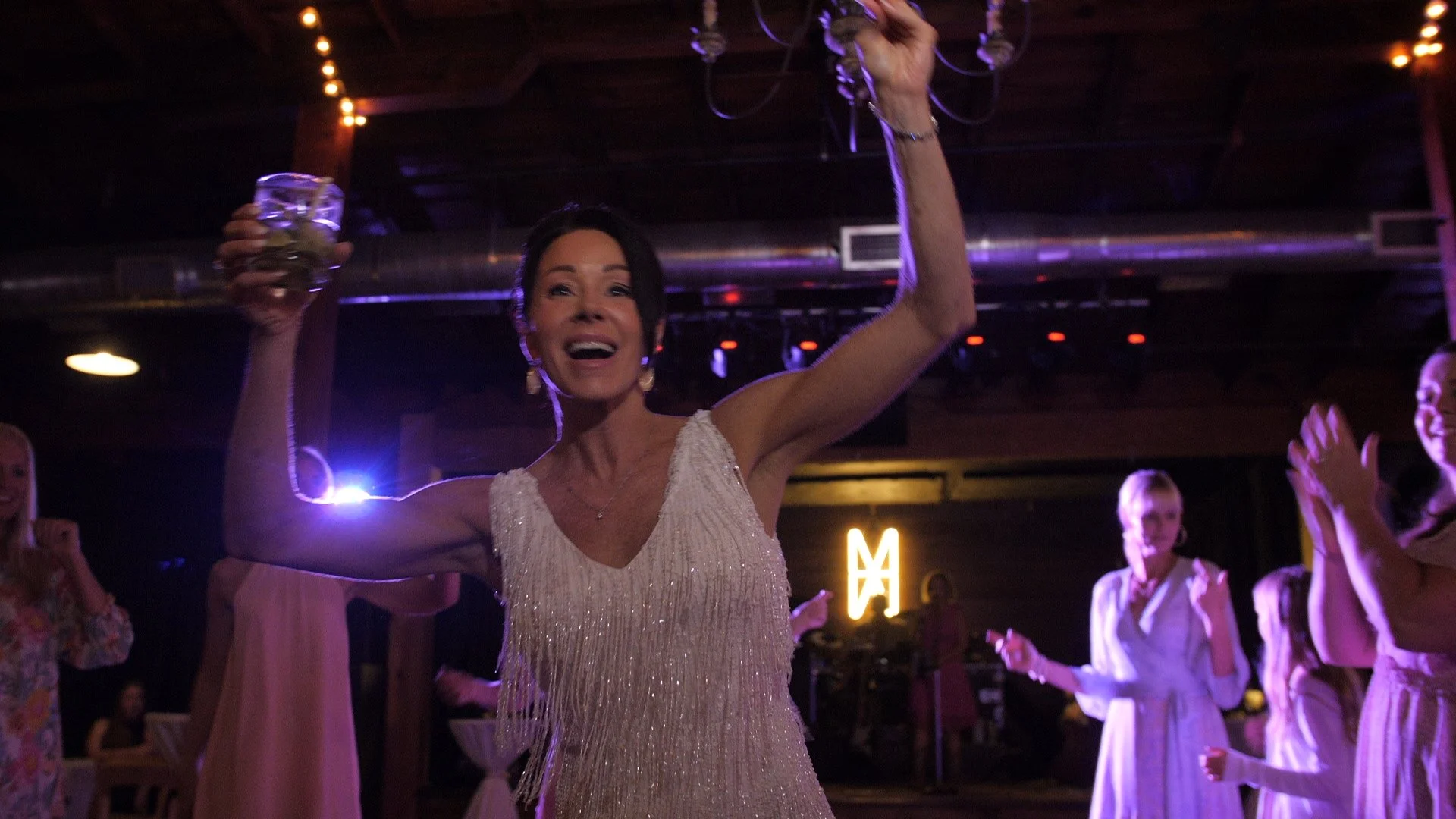 Woman in a glamorous dress celebrating with a drink in a lively party or wedding reception setting, surrounded by other guests.