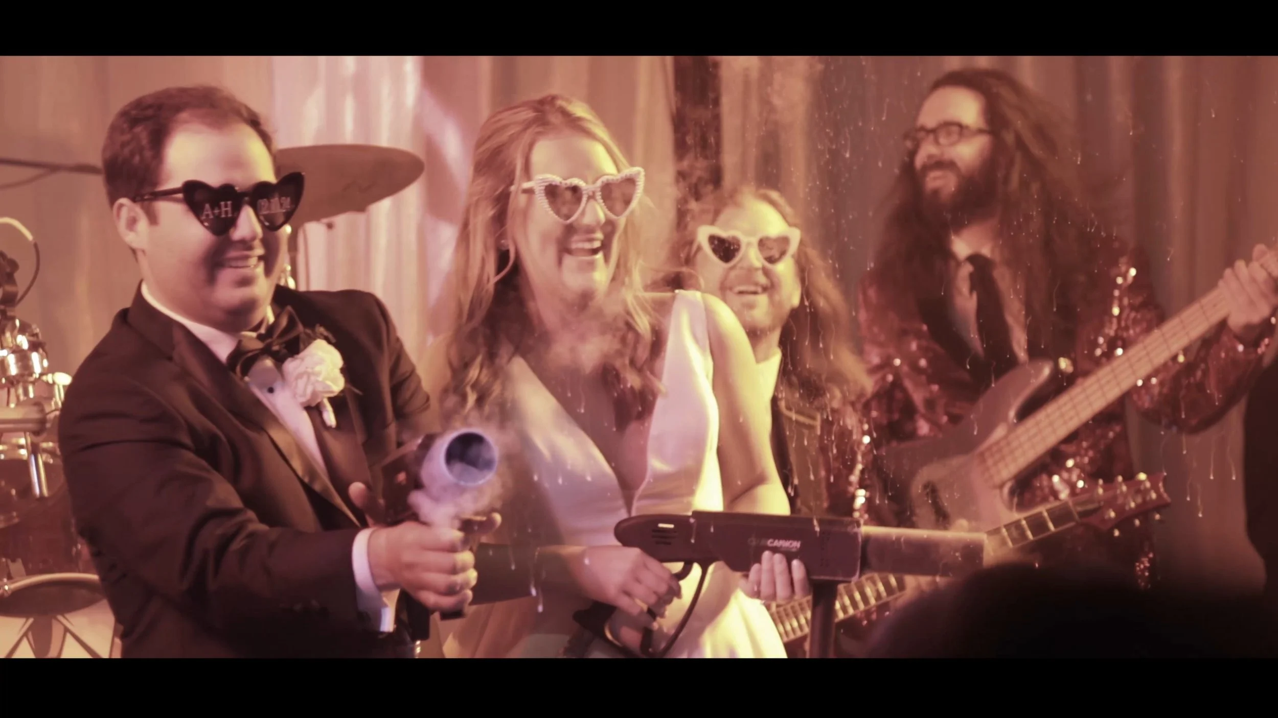 A group of four people at a party, wearing sunglasses, standing close together, with two holding drinks and one playing a guitar, in a lively, decorated setting.