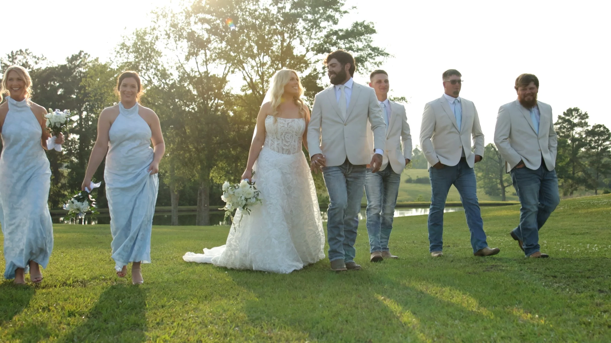 A group of people walking outdoors on a grassy field during a wedding, with women in white dresses and men in white jackets and casual jeans, holding bouquets, with trees and sunlight in the background.