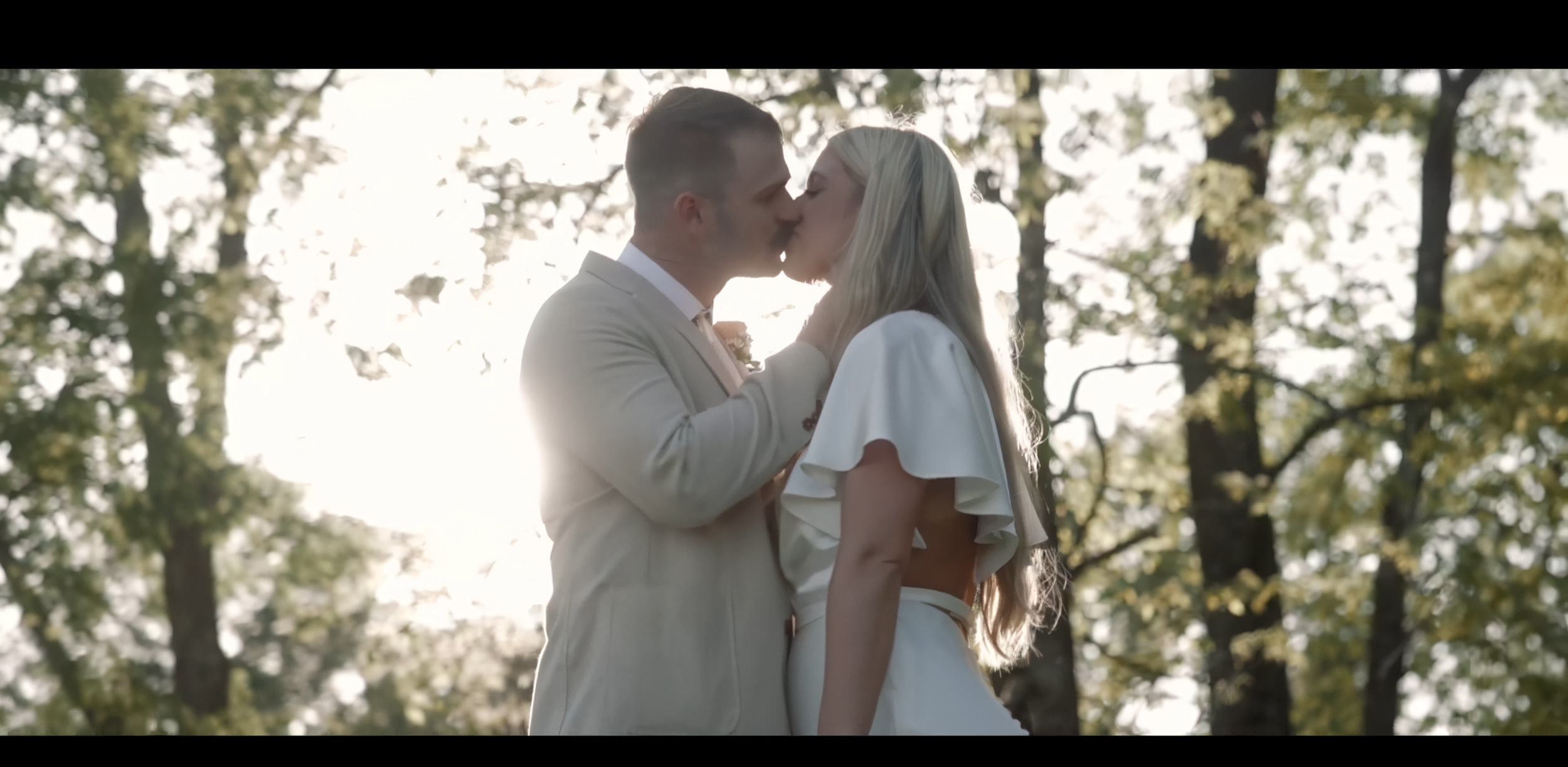 A man and woman share a kiss outdoors during daytime, surrounded by trees with sunlight filtering through leaves.