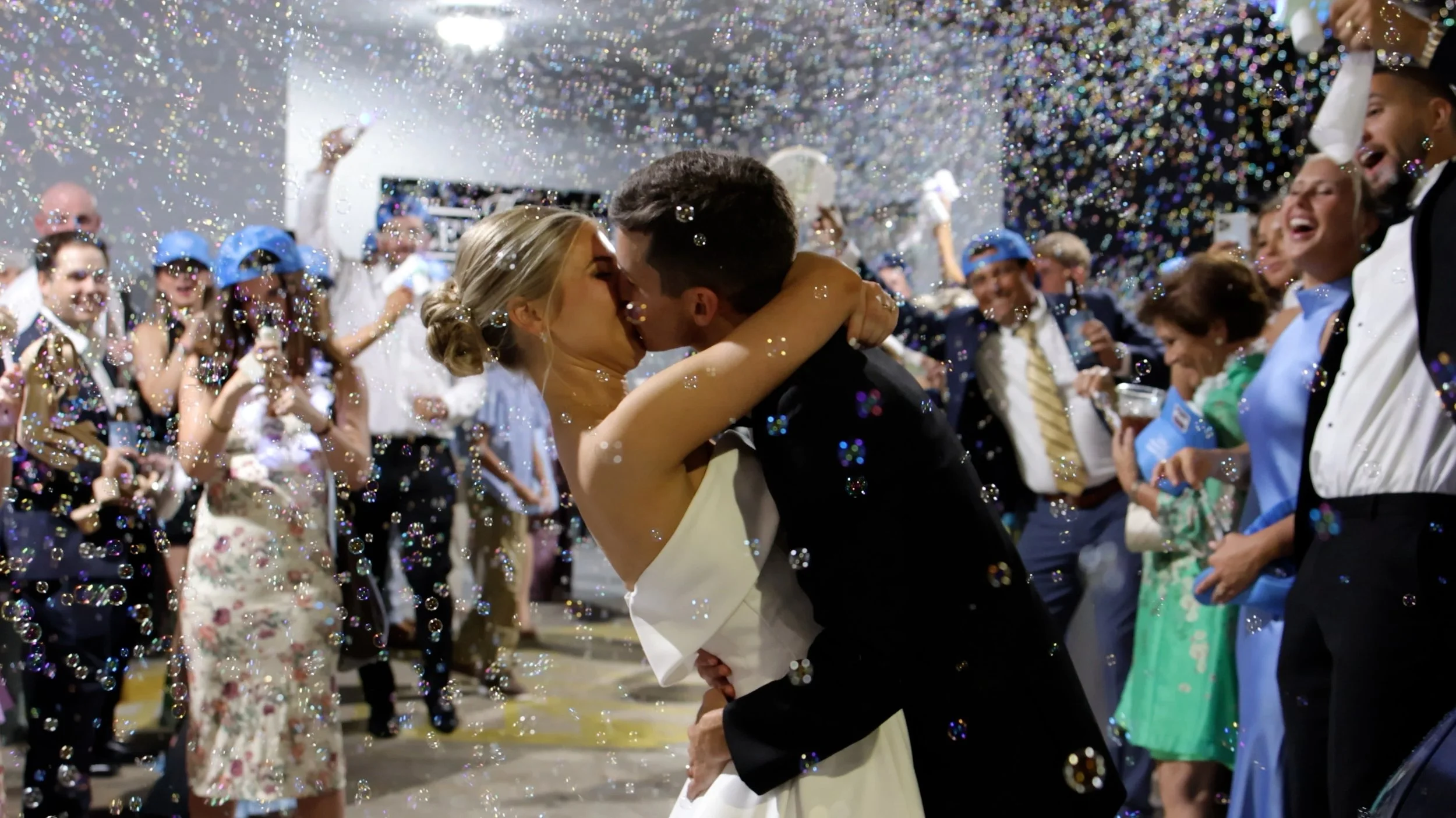 A newlywed couple is kissing at their wedding reception, surrounded by guests and confetti in the air. Louisiana based wedding videography