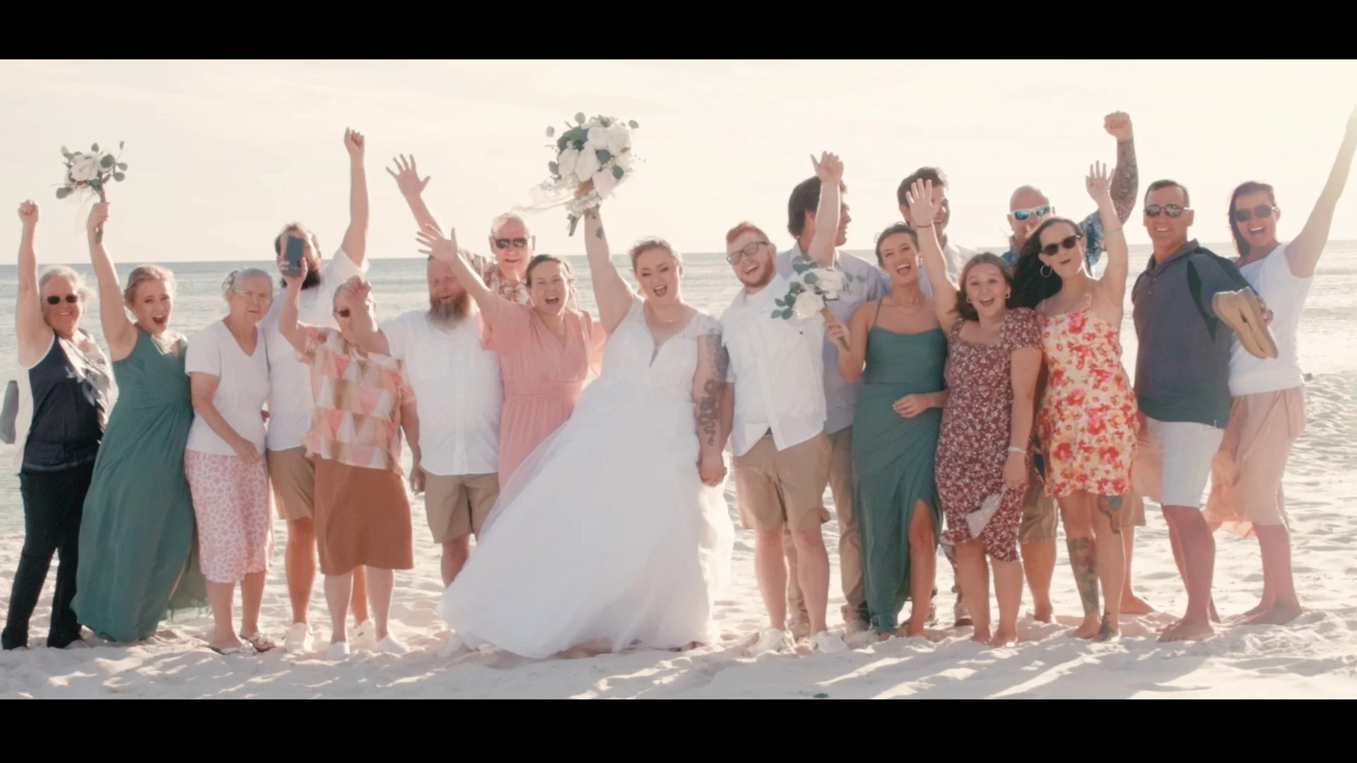A group of people on the beach celebrating a wedding, including the bride holding hands with the groom, surrounded by friends and family.
