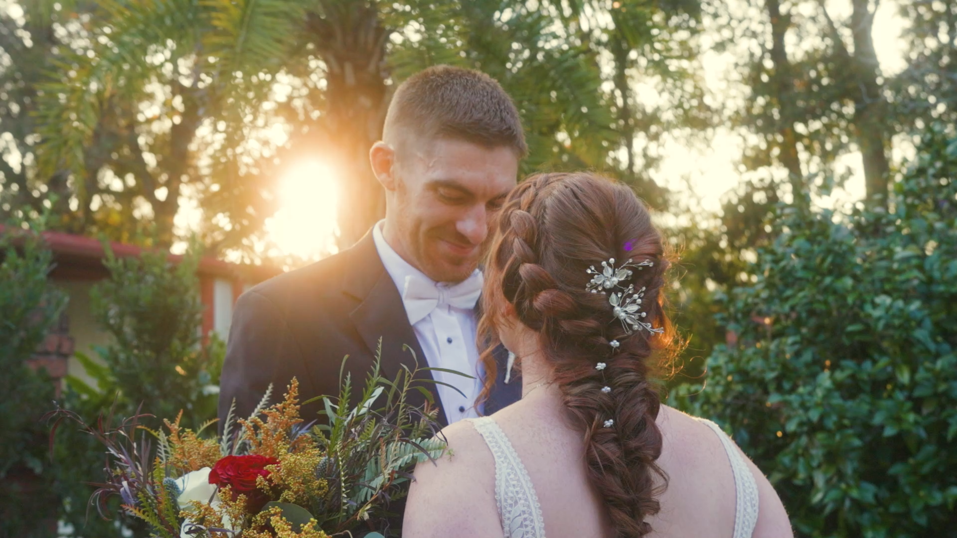 A bride and groom share a kiss outdoors during sunset, surrounded by green foliage, with the bride holding a bouquet of flowers.