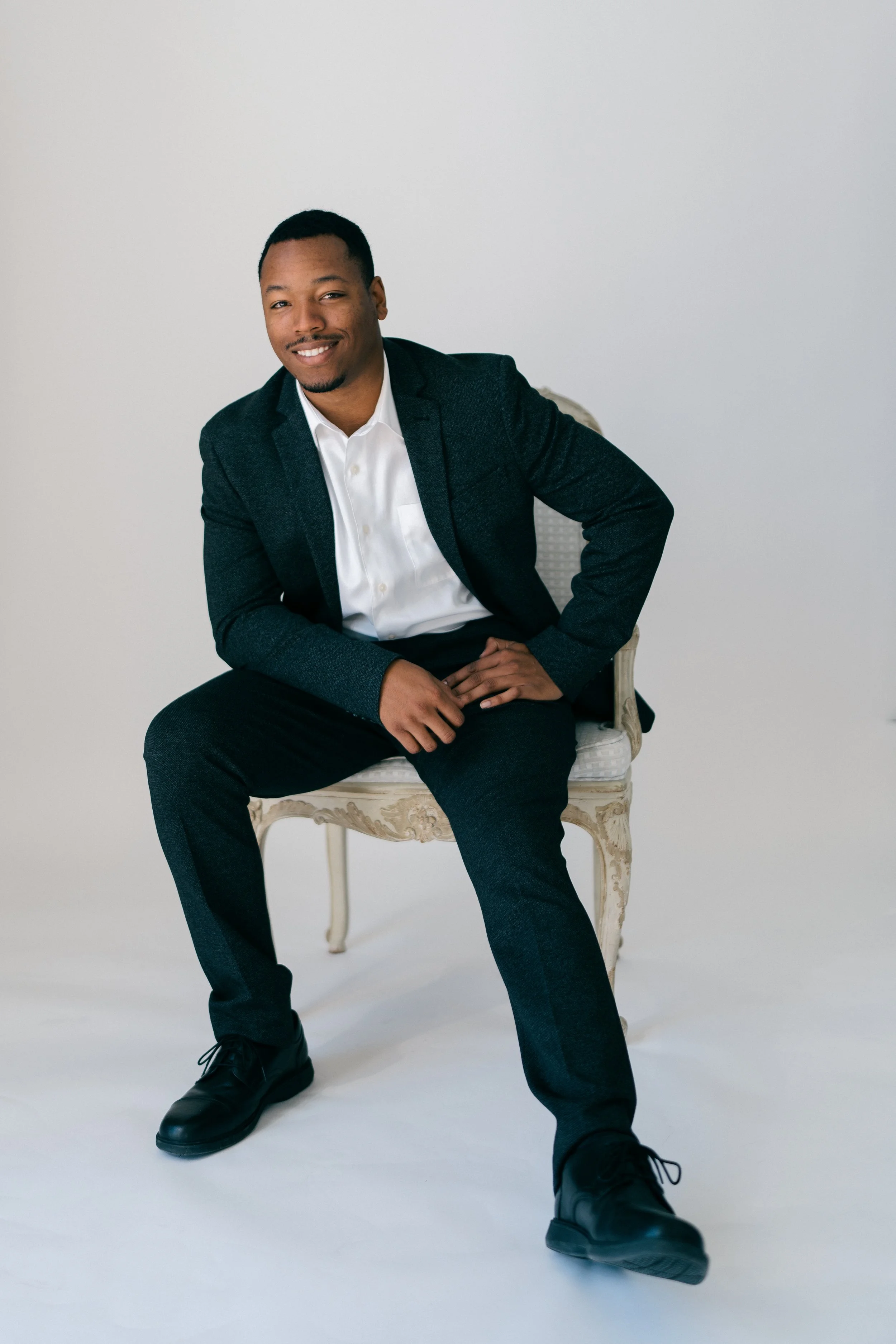 Colby Glaude, owner of Glaude Videography in a dark blazer, white shirt, and dark pants sitting on a decorative white chair against a plain light background.