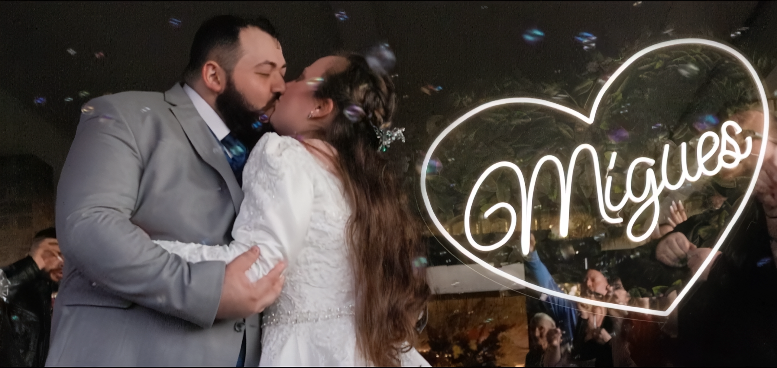 A newlywed couple sharing a kiss at night, with a large illuminated heart-shaped sign that reads 'Mr & Mrs' behind them, reflecting in a mirror.
