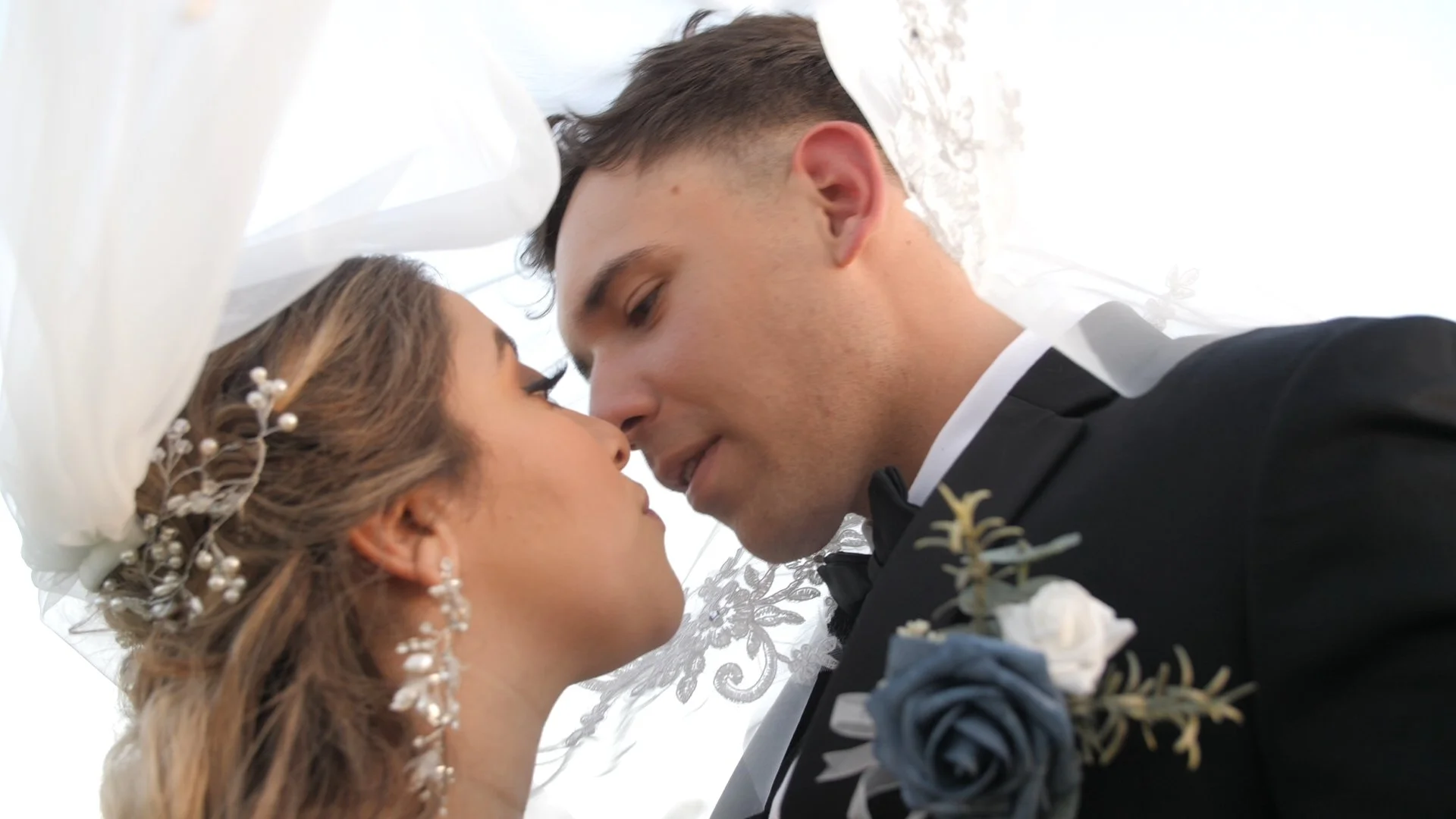 A bride and groom are close together, with their foreheads touching, under a white lace umbrella during their wedding celebration.