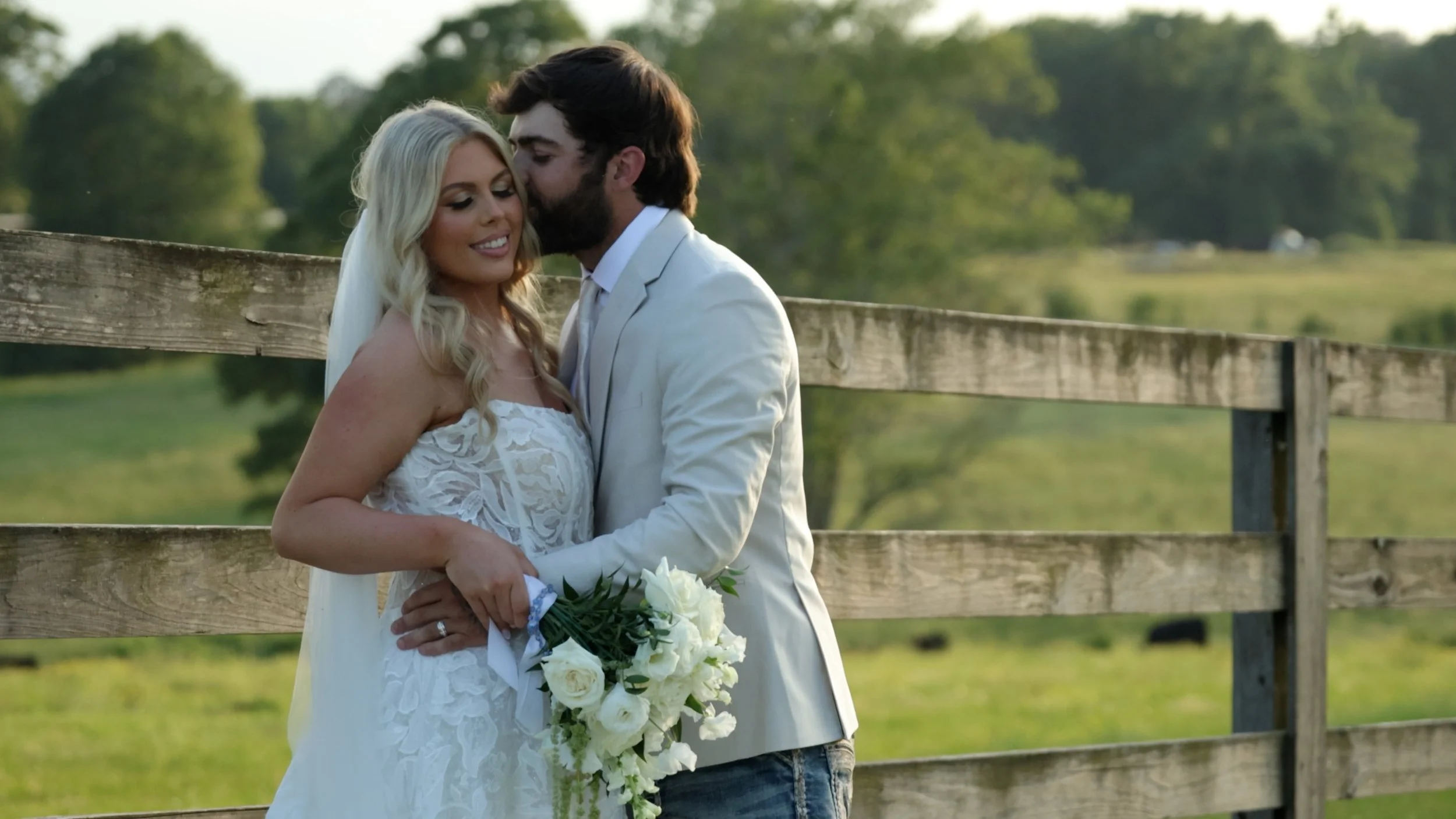 A bride and groom embrace outdoors near a wooden fence, with greenery in the background, during a wedding.