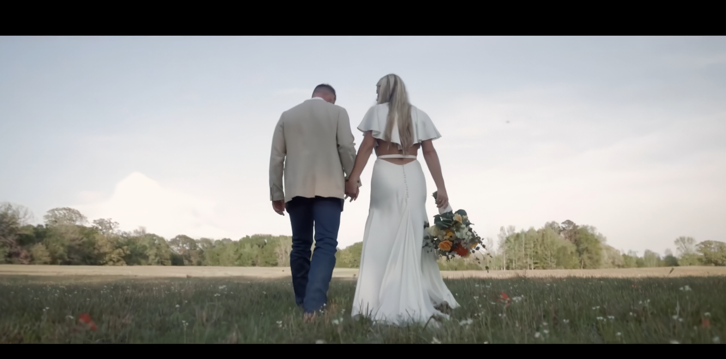 A couple walking hand in hand outdoors during their wedding, with the bride holding a bouquet of flowers, in a grassy field with trees in the background under a cloudy sky.