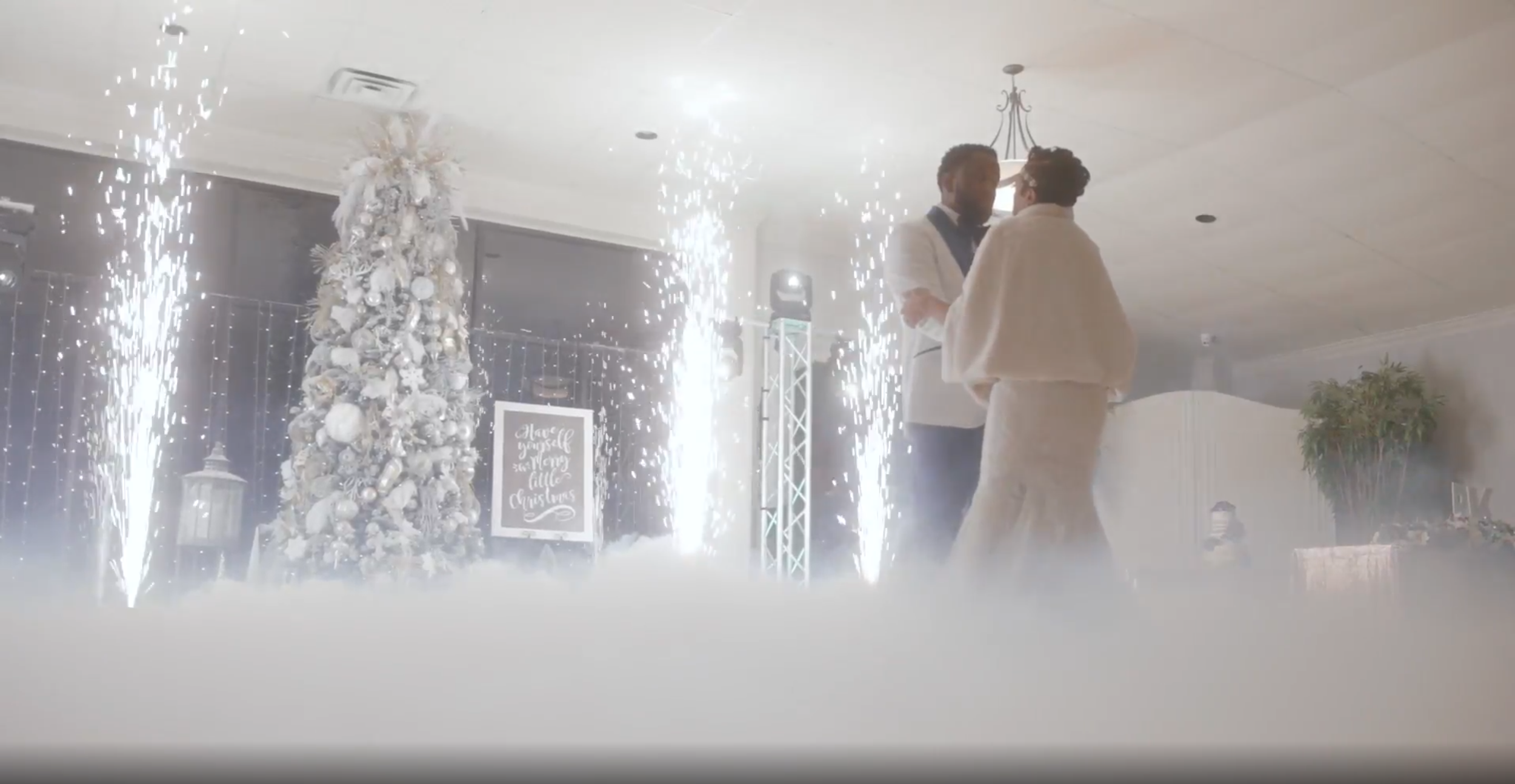 A couple dancing at a wedding reception with a decorated Christmas tree, a sign saying 'Have yourself a merry little Christmas,' and fireworks display in the background.