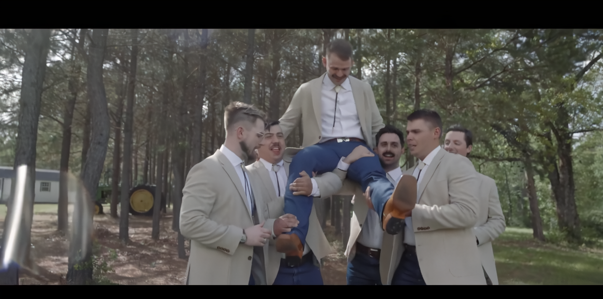 A group of men in beige suits lifting a man in a beige jacket onto a wooden rail in a wooded outdoor area.