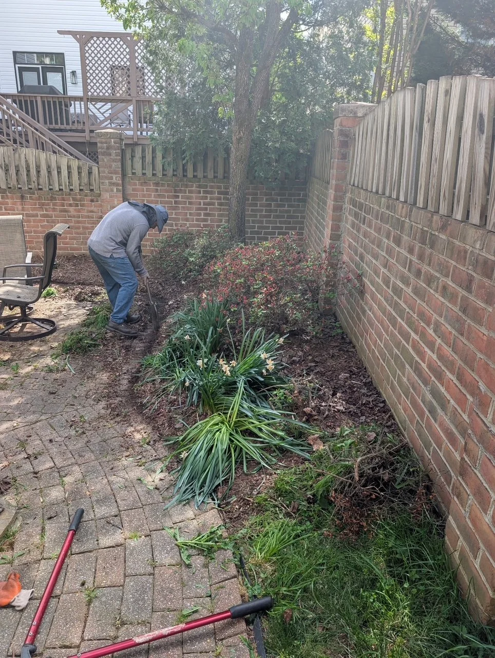 Person gardening in a backyard, trimming bushes along a brick wall with garden tools, on a paved patio with furniture.