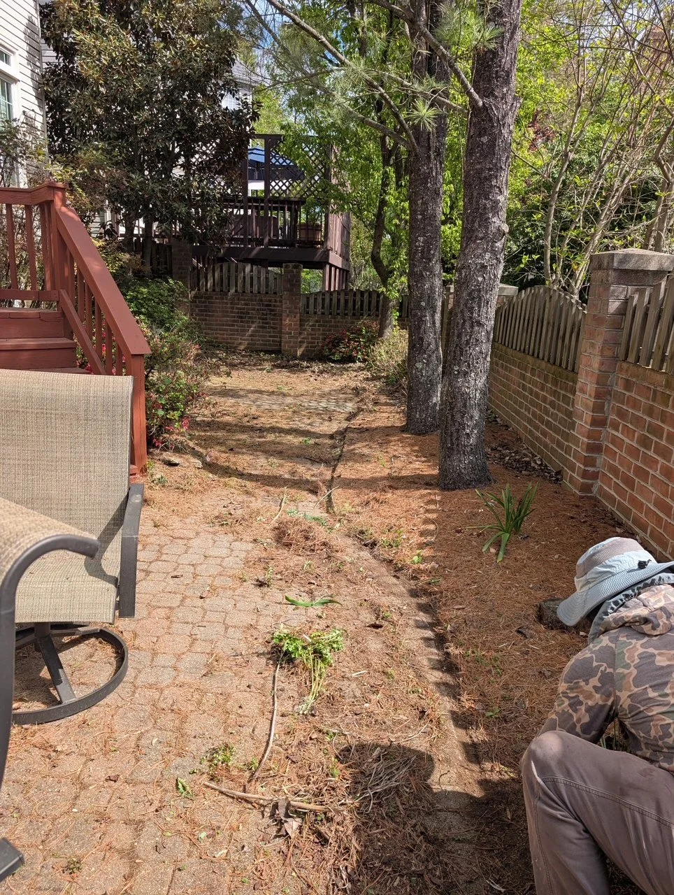 Person in camouflage hoodie and white hat working in a backyard garden with trees, a brick wall, and a wooden deck in the background.