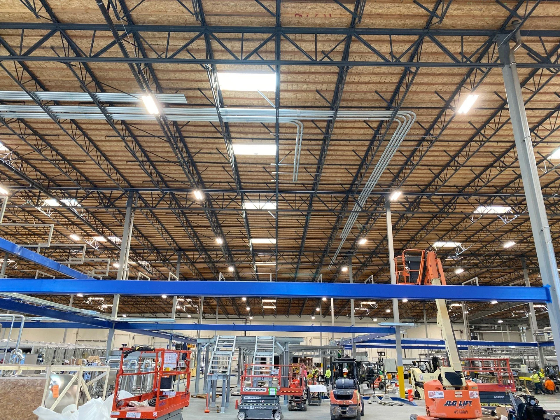 The interior of a large warehouse or industrial building under construction, with visible metal framing, ceiling insulation, and workers operating equipment.