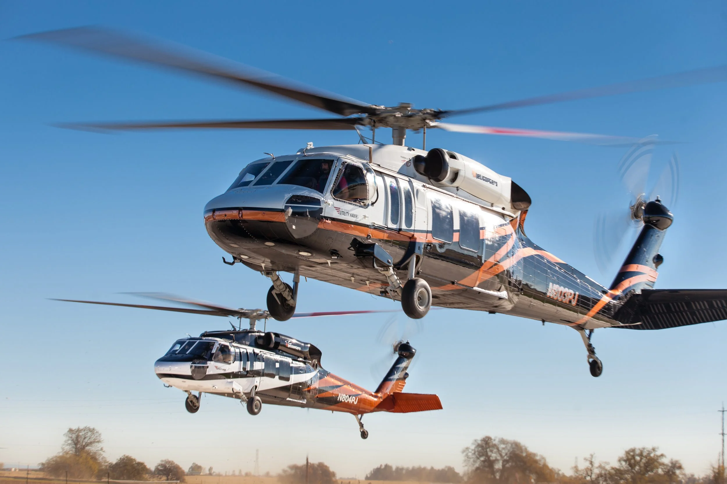 Two helicopters flying in formation against a clear blue sky over a rural landscape.