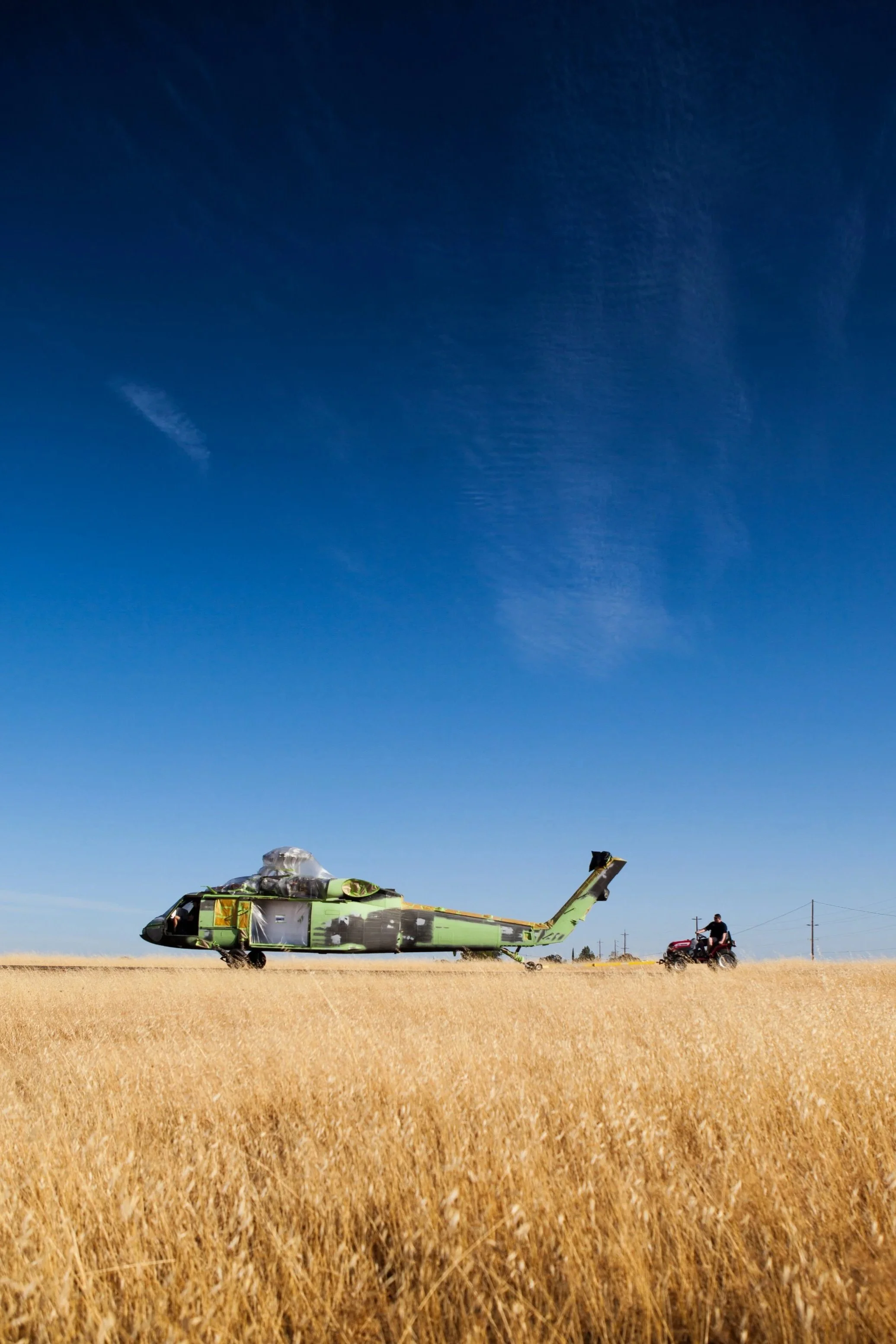 A helicopter and a person on a motorcycle in a golden wheat field under a clear blue sky. The helicopter is painted in shades of green and yellow and appears to be a military or rescue type, with some equipment or cargo covered on top.