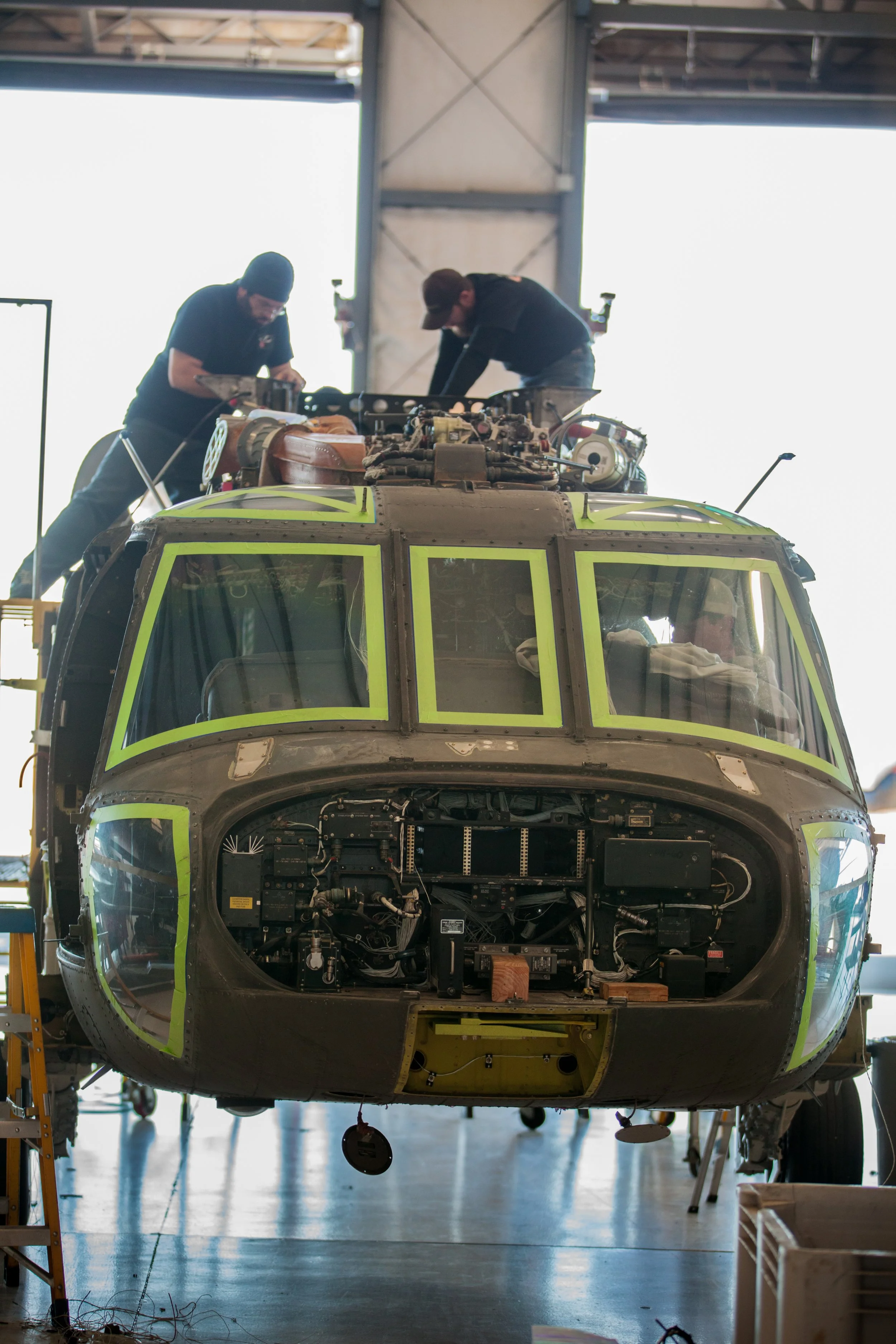 Two technicians working on the top of a helicopter inside a hangar, with its cockpit and internal components exposed.