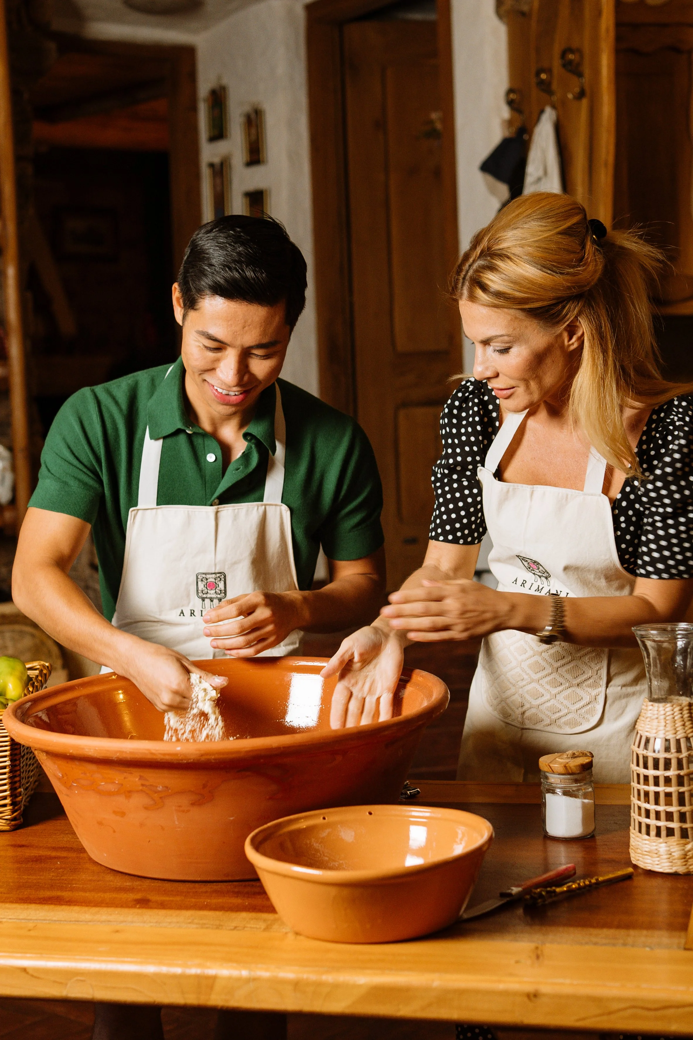 A man and a woman in a rustic kitchen, both wearing aprons, are baking together with various bowls and utensils on the table in front of them.