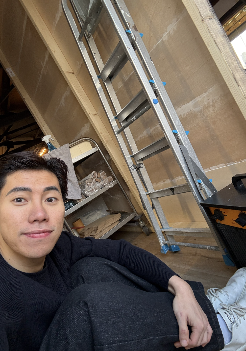 A young man taking a selfie in a construction or renovation site with a stepladder, a metal cart with glassware, and wooden wall panels in the background.