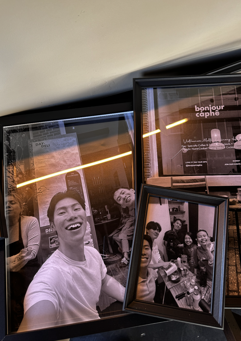 A wall display featuring two framed photographs of a group of friends smiling and having fun indoors, with reflections of neon lighting and a menu on the glass.