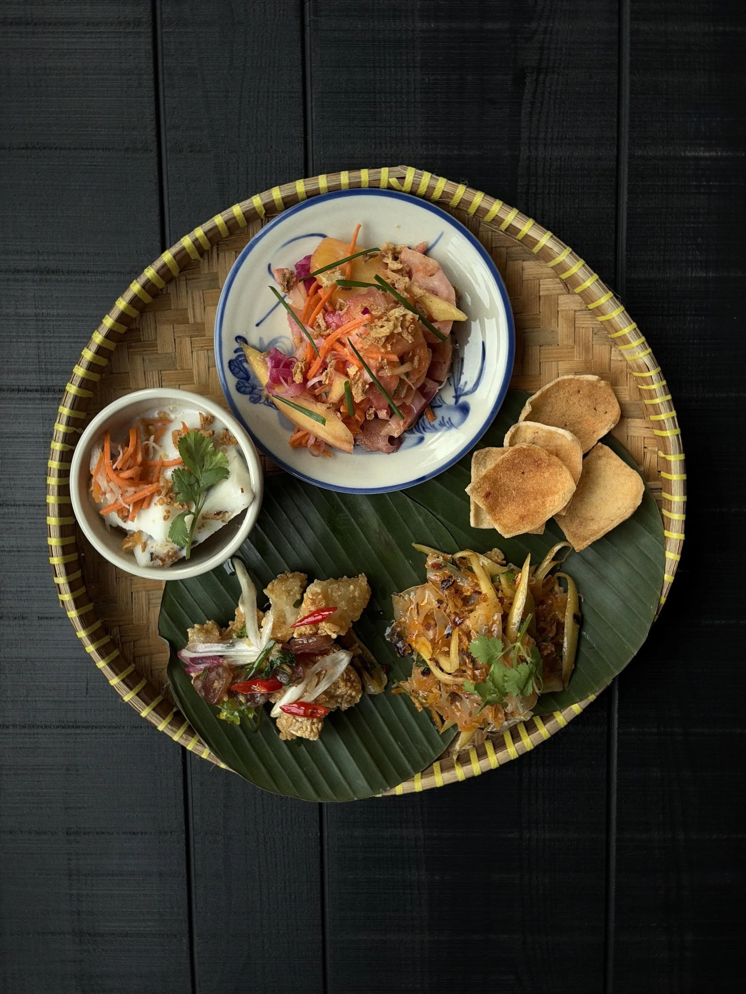Assorted Asian appetizers served on a woven tray with banana leaf, including fresh vegetables, fried spring rolls, and a small bowl of dipping sauce garnished with cilantro.