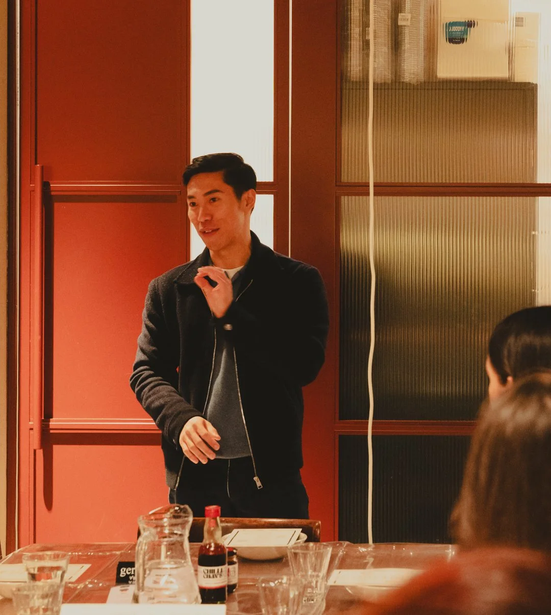 A man speaking at a dinner gathering, standing by a table with drinks and condiments, inside a warmly lit room with wooden paneling and patterned glass panels.