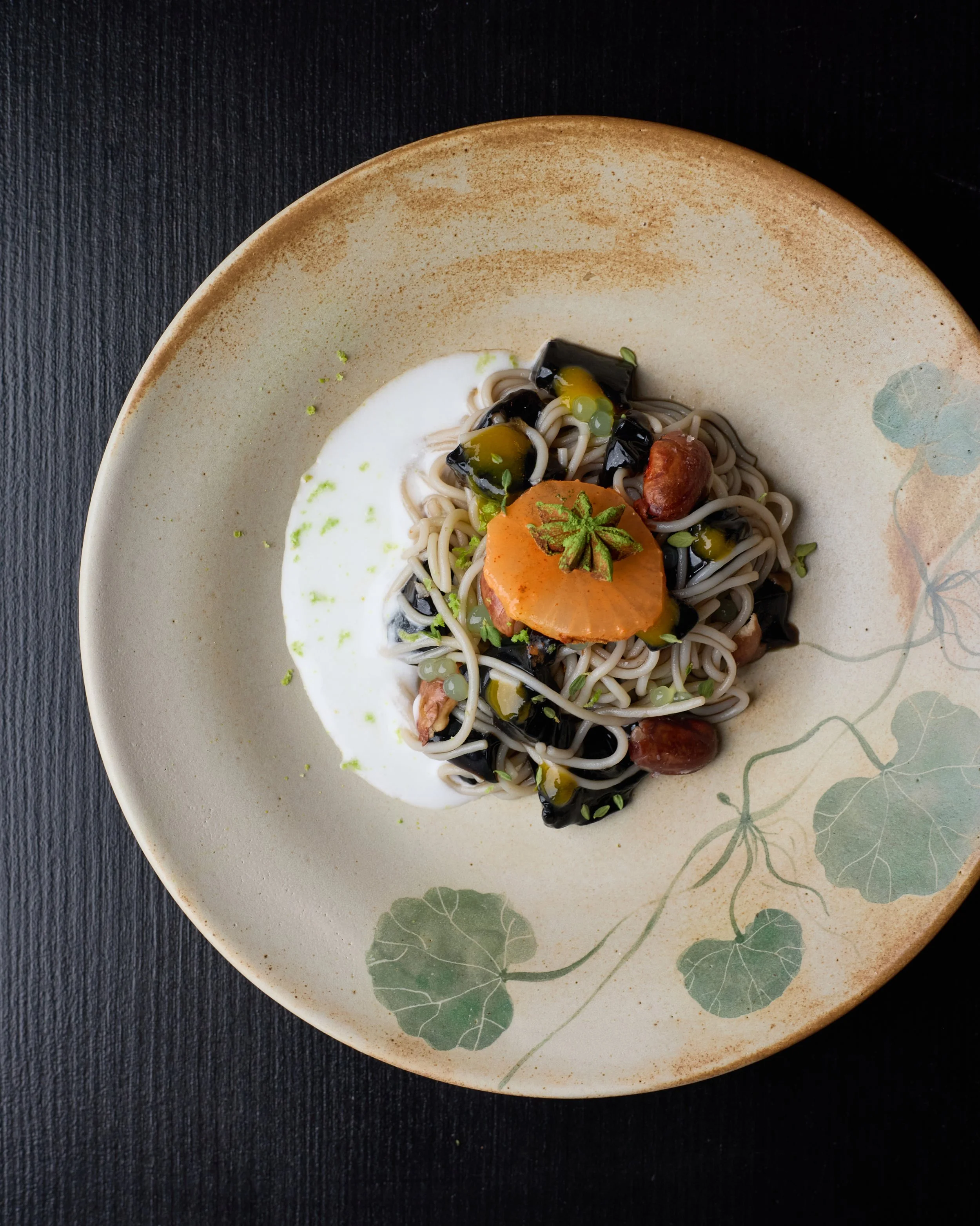A ceramic plate with a floral pattern holding soba noodles topped with vegetables and garnishes, placed on a dark surface.