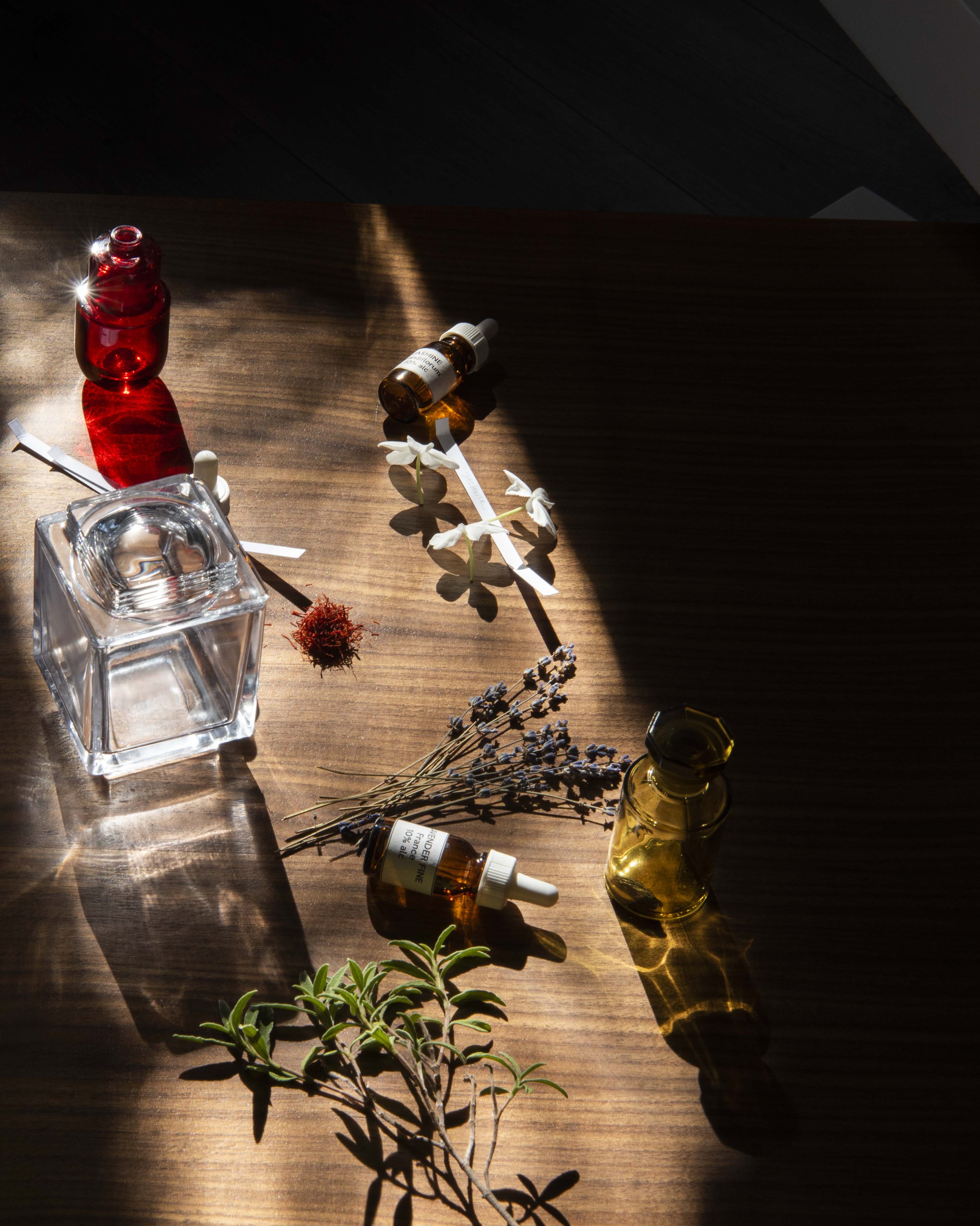 A wooden surface with sunlight and shadows. On it are a small red glass bottle, two amber essential oil bottles, a clear glass ashtray, a sprig of green herb, and flowers including lavender, white orchids, and a small dried red flower.