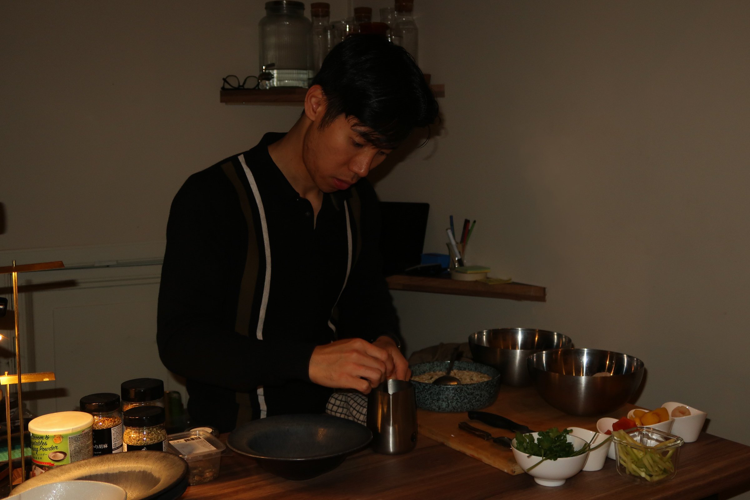 A person with dark hair preparing food in a kitchen, surrounded by bowls of ingredients and spices, standing at a wooden counter.