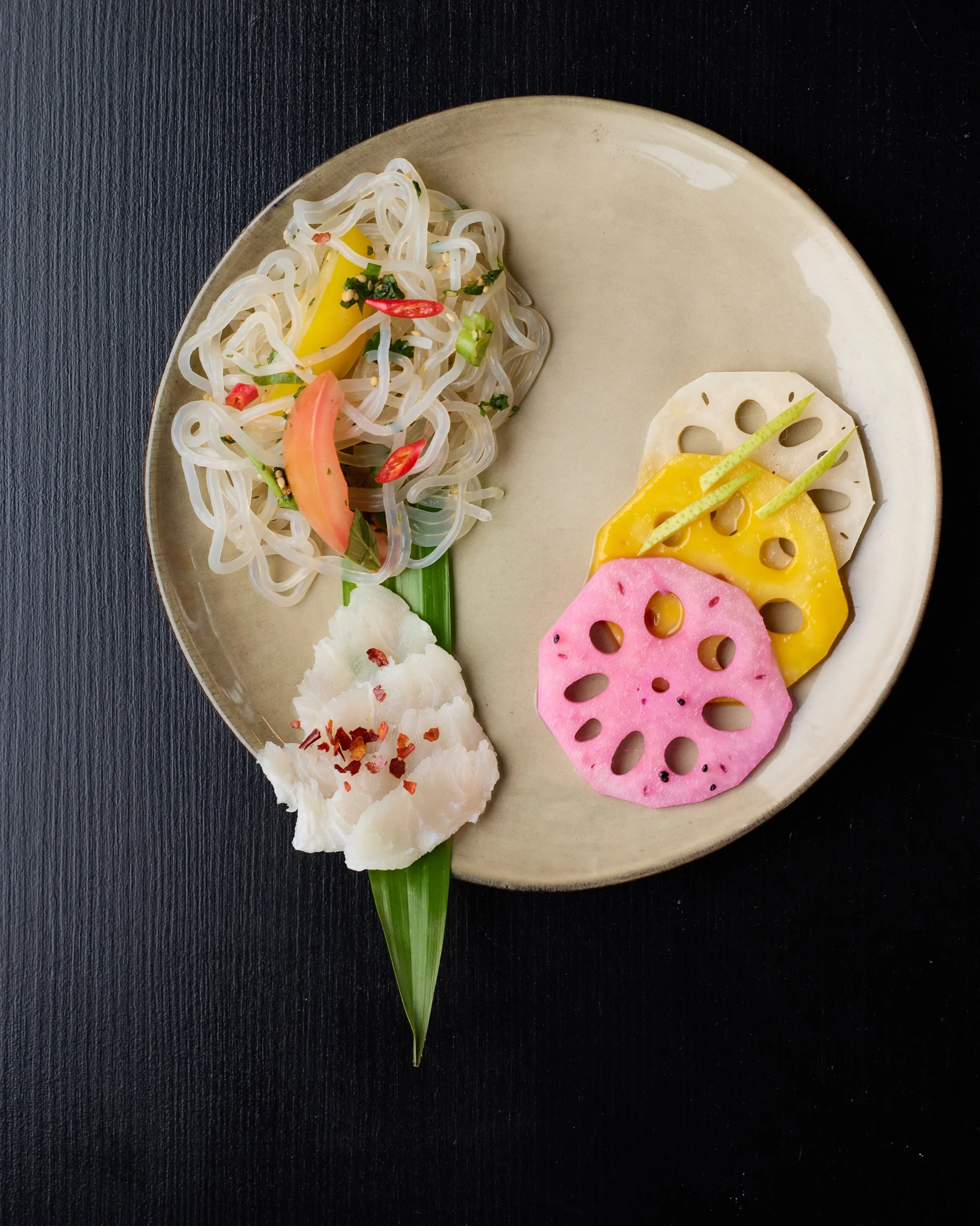 Plate of assorted Asian-style appetizers including pickled vegetables, lotus root slices, and rice noodles with garnishes on black background.
