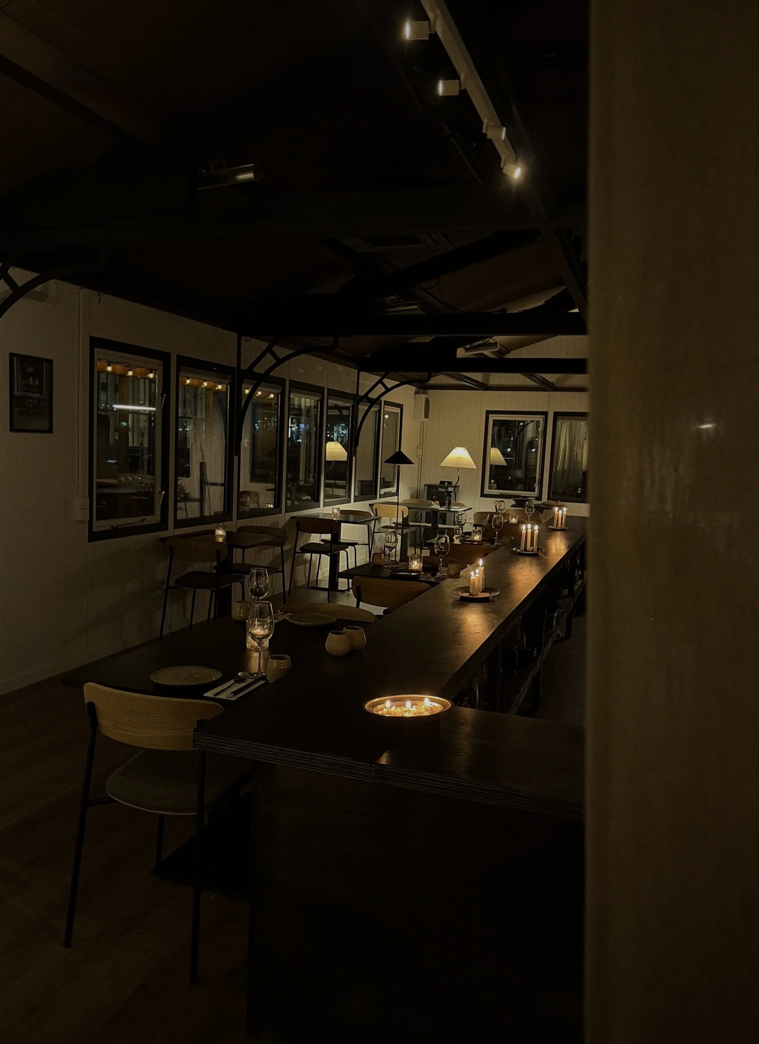 A dimly lit restaurant interior with tables set for dining, candles on tables, and warm lighting from lamps on the walls.