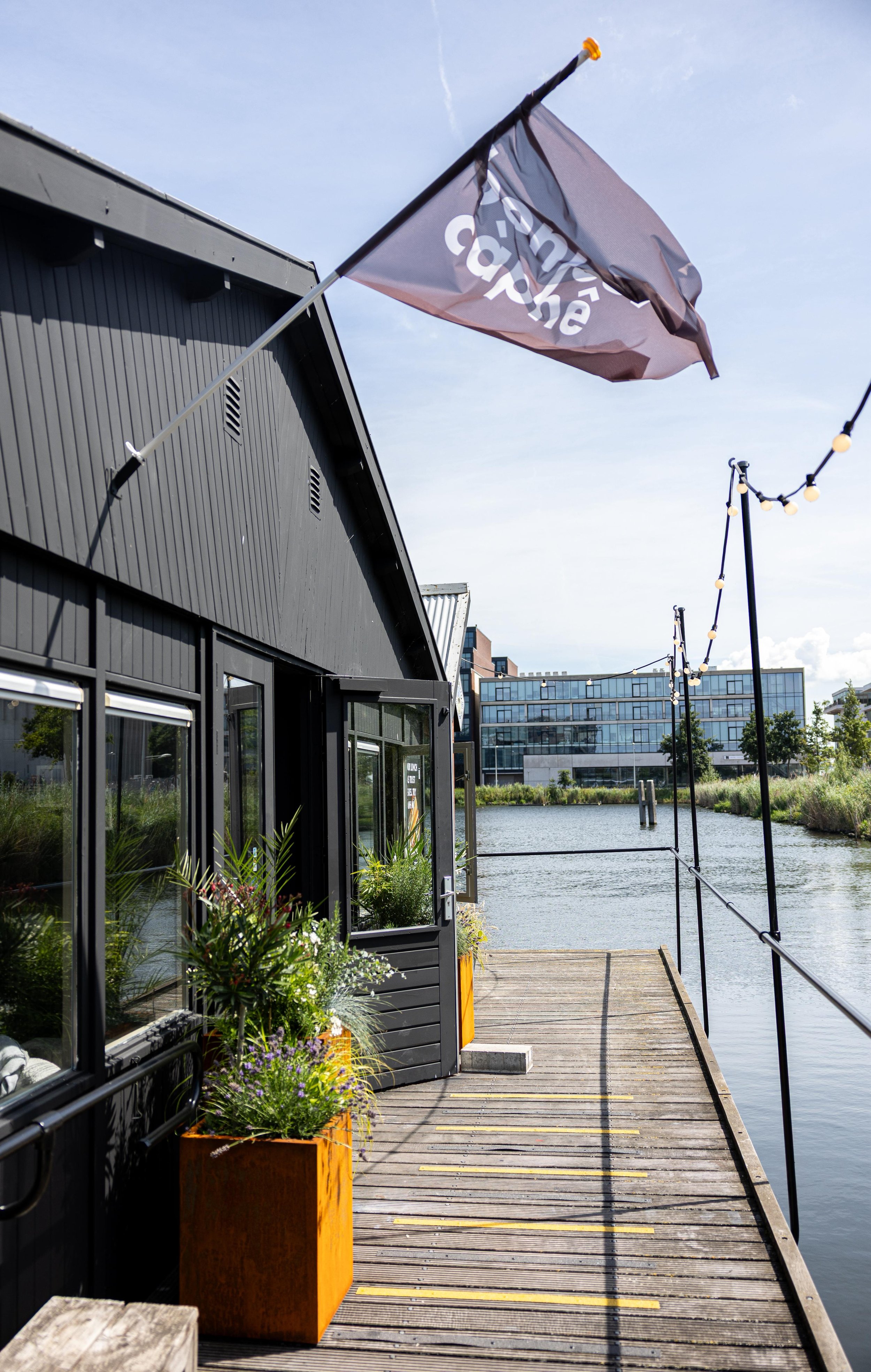 A modern black building next to a body of water with a wooden dock. A flag with white text on a black background is flying on a pole attached to the building. Outdoor string lights are hanging along the water, and there are green plants in bright orange planters near the building. In the background, there are glass buildings and a partly cloudy sky.