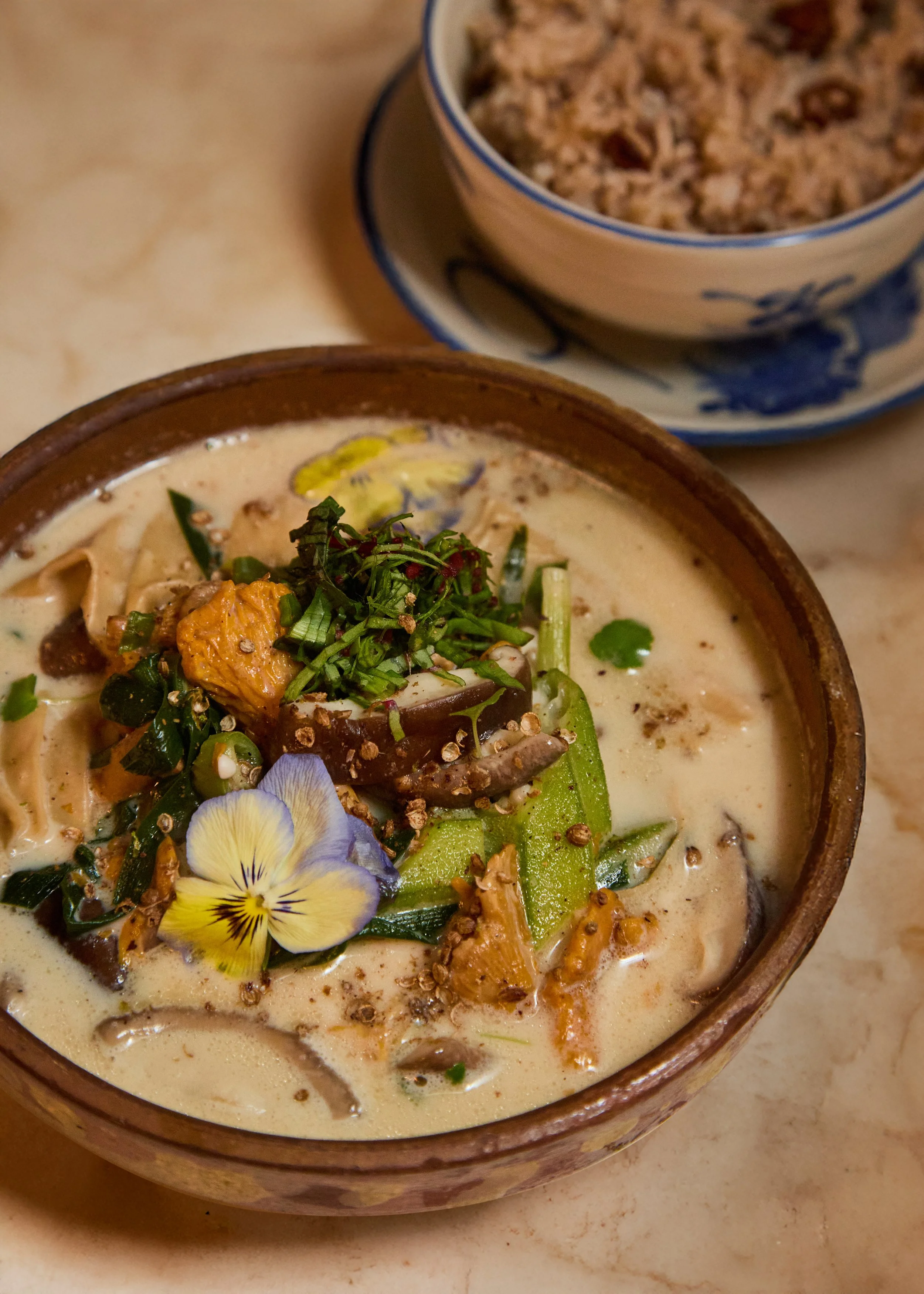 A bowl of Vietnamese creamy soup garnished with edible flowers, chopped herbs, and microgreens, with a side of grain or rice in a smaller bowl behind it.