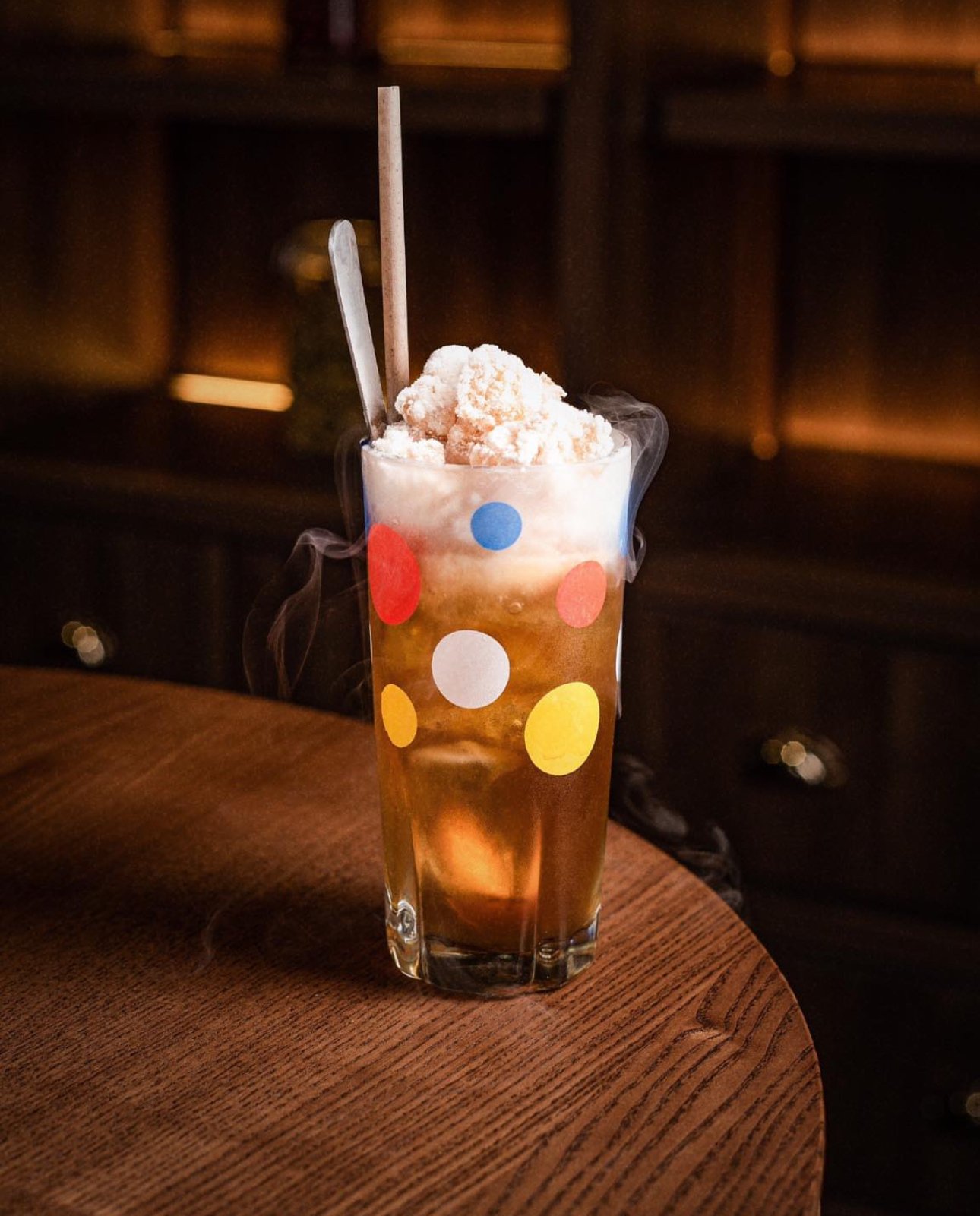 A tall glass filled with a dark-colored beverage, topped with cotton candy, with a metal straw and a pink straw, sitting on a wooden table in a dimly lit environment.