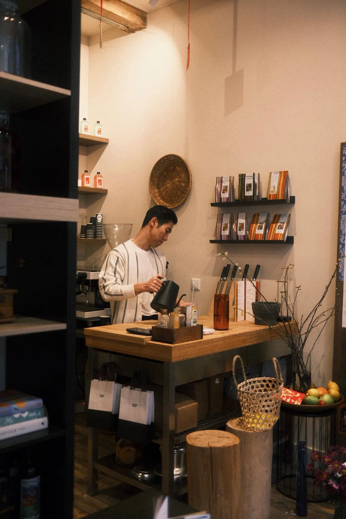 A man preparing Vietnamese coffee behind a wooden counter in a cozy cafe with shelves of books, a woven basket on the wall, and fresh fruits on a nearby table in Amsterdam.