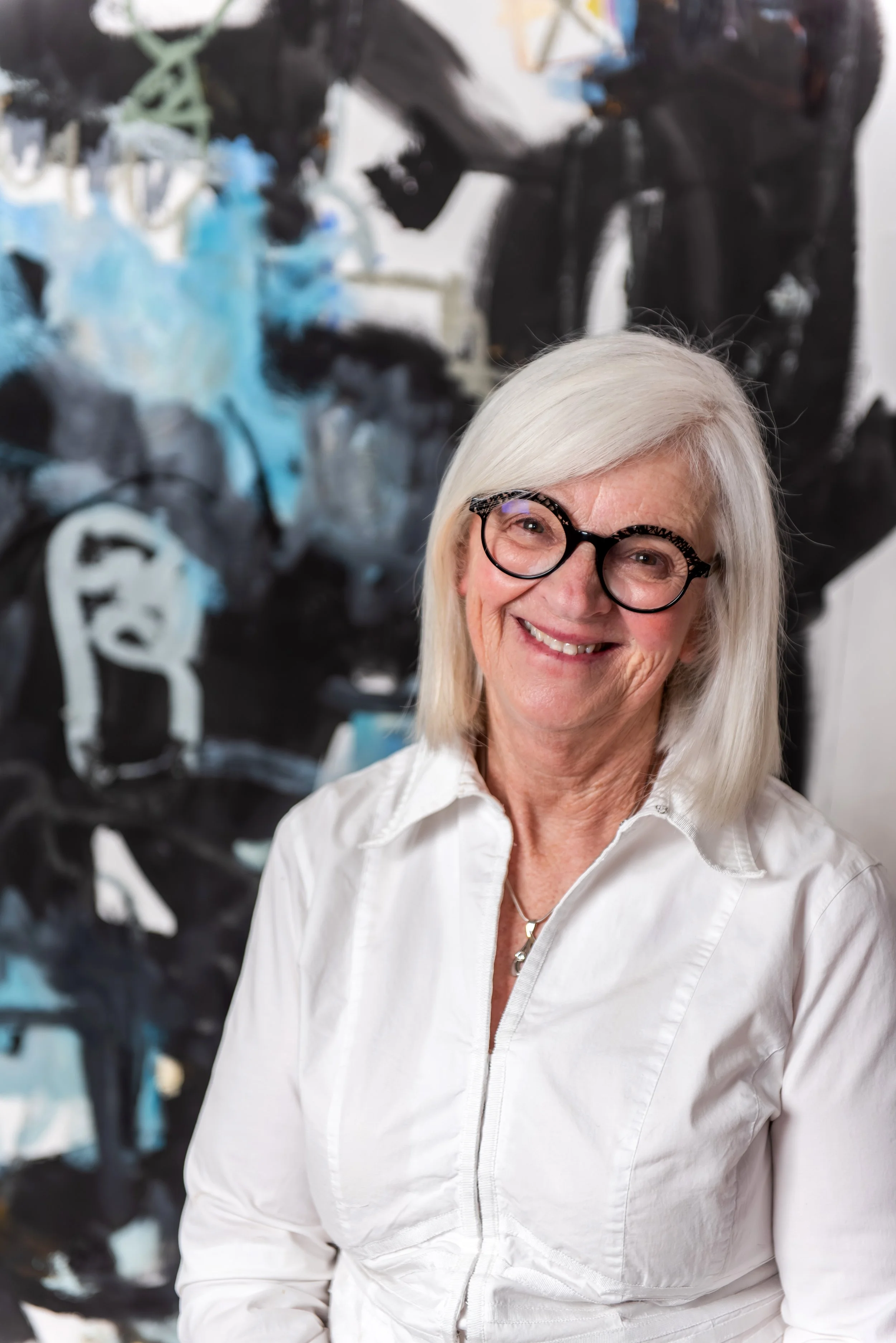 Smiling woman with long white hair, wearing glasses and a white button-up shirt, standing in front of a painting with dark and light abstract patterns.