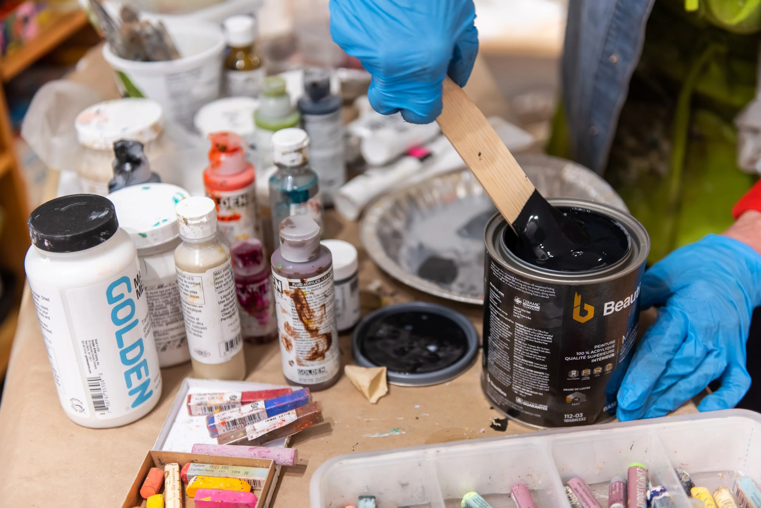 Artist's workspace with bottles of paint, chalk pastels, and paints, with a person in blue gloves holding a wooden stick in a can of black paint.