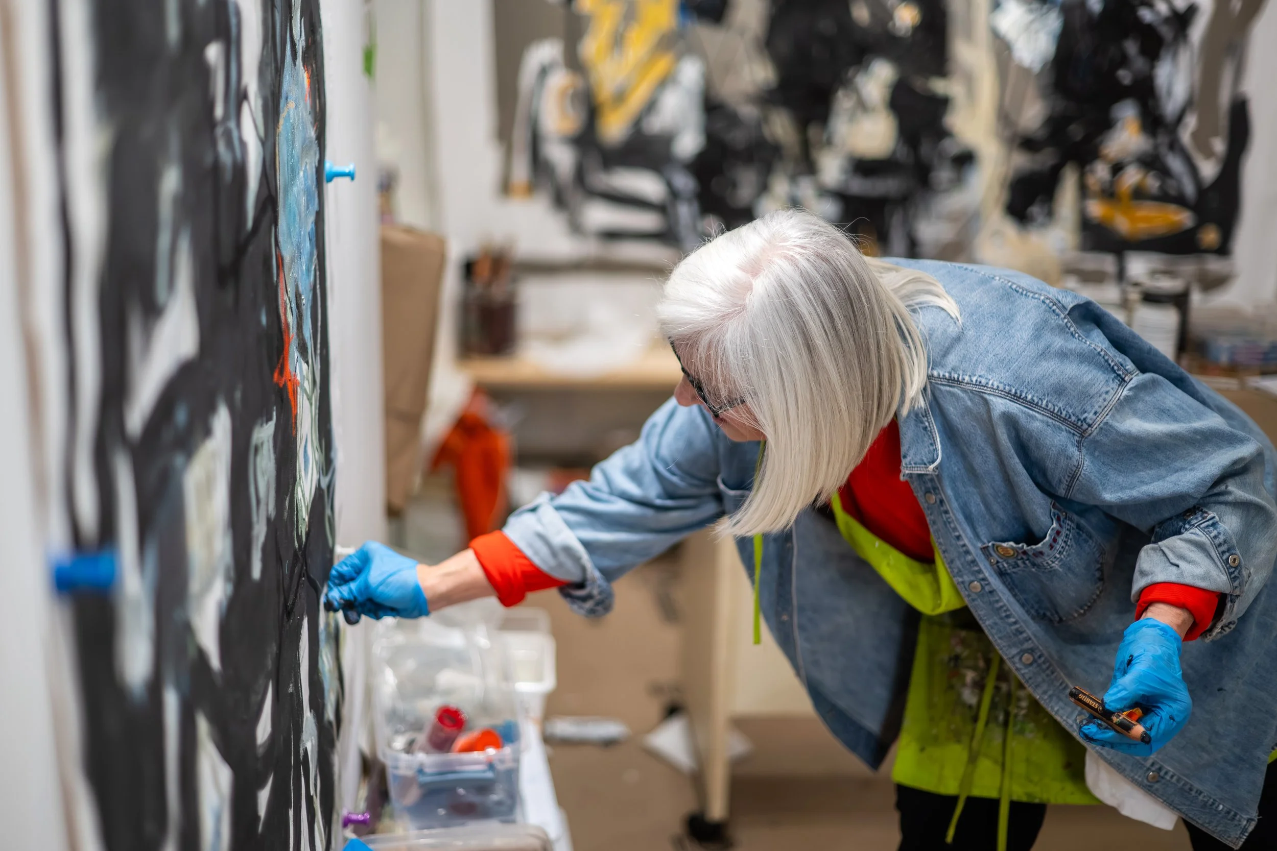 A woman with gray hair, glasses, a denim jacket, and blue gloves is working on a black, white, and orange abstract painting in an art studio.