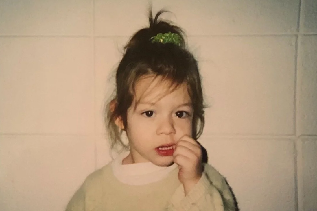 Young girl with light brown hair tied up with a green hair clip, standing against a beige tiled wall, wearing a beige sweater, touching her nose with her hand.