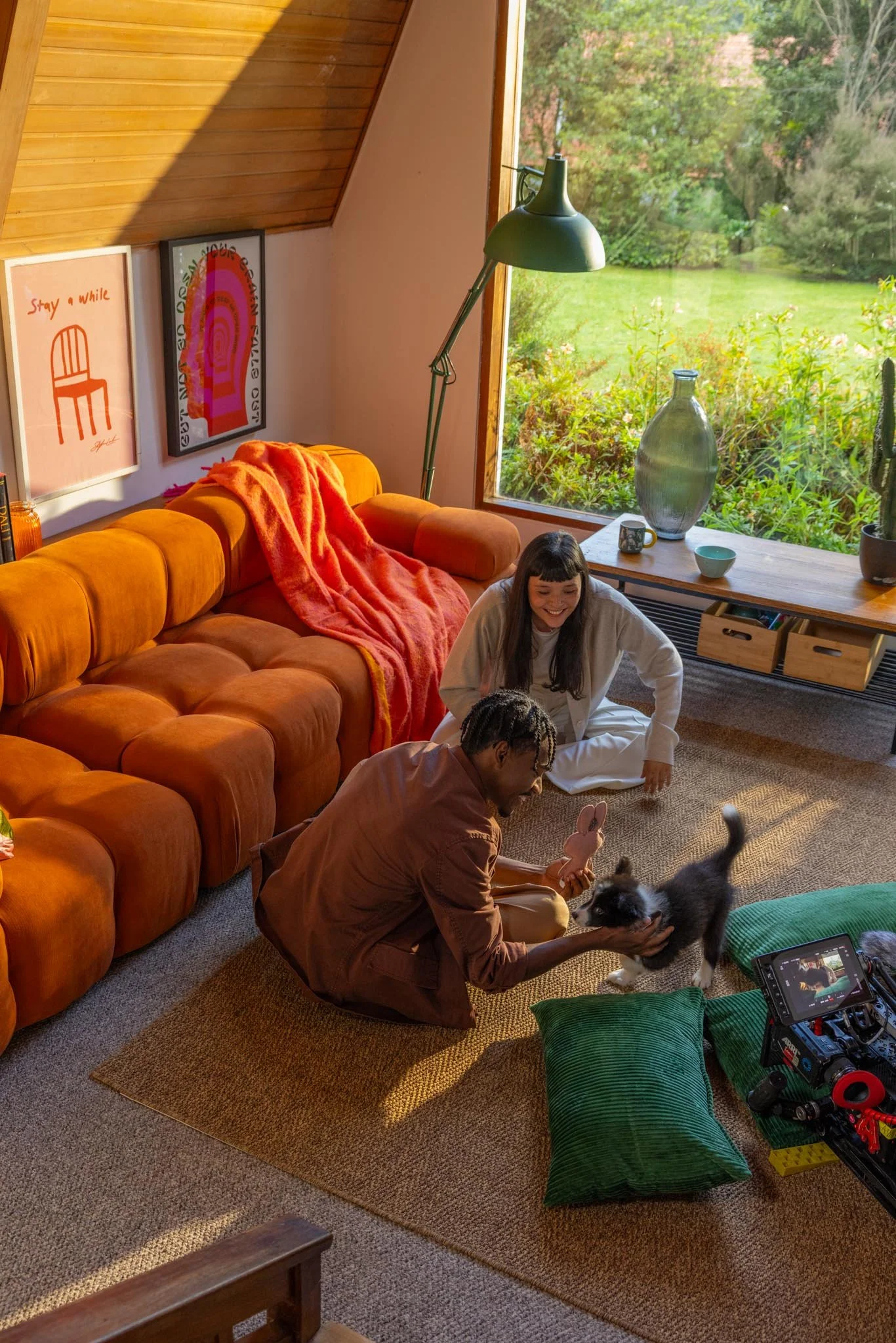 A cozy living room with a large orange couch, a woman with dark hair smiling, man holding a puppy, and a puppy in the foreground. Large window with greenery outside, wall art, a green desk lamp, a large glass vase, and various small decorative items 